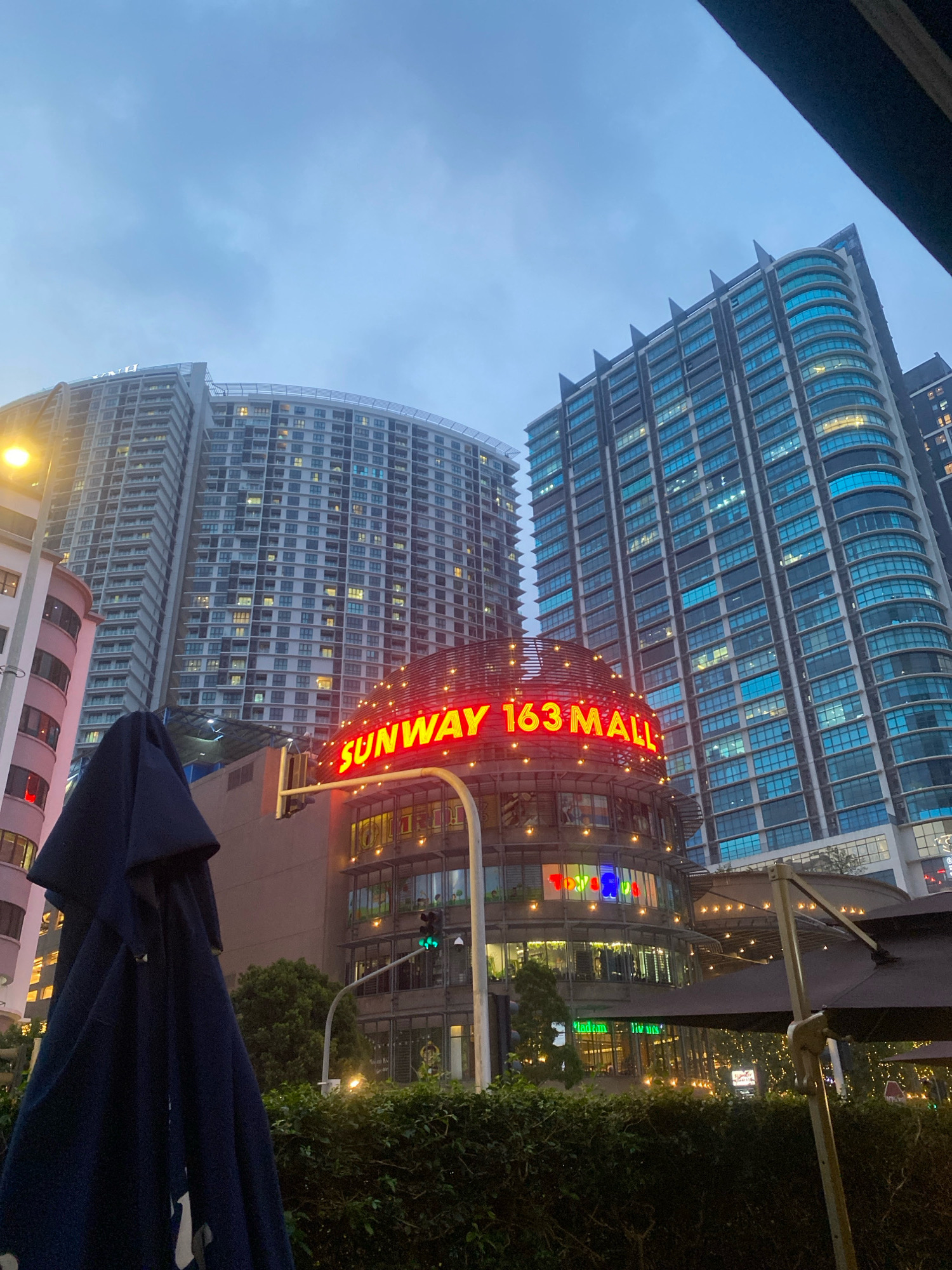 The image depicts a nighttime scene in Kuala Lumpur, Malaysia, featuring a modern cityscape with prominent architecture and signage. In the foreground, a dark blue umbrella, partially furled, dominates the left side of the frame. Behind it, a lush green hedge runs along the bottom. To the right, part of a dark-colored umbrella with a metal pole is visible, casting a shadow.
The central focus of the image is the "SUNWAY 163 MALL" sign, illuminated in bright red neon letters against a rounded, multi-story building. The mall structure itself is adorned with decorative string lights. Adjacent to the mall, two towering modern buildings rise into the evening sky. The building on the right is characterized by its blue-tinted glass facade, reflecting the dim light. The building on the left appears to be residential, with numerous windows illuminated by interior lights.
A traffic light showing a green signal is visible on the mall's facade, indicating ongoing traffic flow. The sky above is overcast, contributing to the twilight atmosphere. The overall scene suggests an urban commercial district, likely a popular spot for shopping and dining, as evidenced by the illuminated mall and surrounding structures. The presence of the umbrella and hedges hints at an outdoor seating area, possibly for a restaurant or cafe.