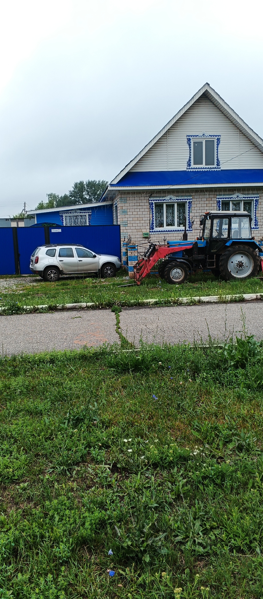 In the village of Podgornoye, Russia, a light-colored Dacia Duster is parked next to a blue metal fence. Adjacent to the Dacia and the fence is a brick house with blue trim and decorative blue window surrounds. In front of the house, a blue tractor with a red front loader is parked on the grass. The scene is set against a cloudy sky and features a paved path in the foreground with overgrown green grass on either side.