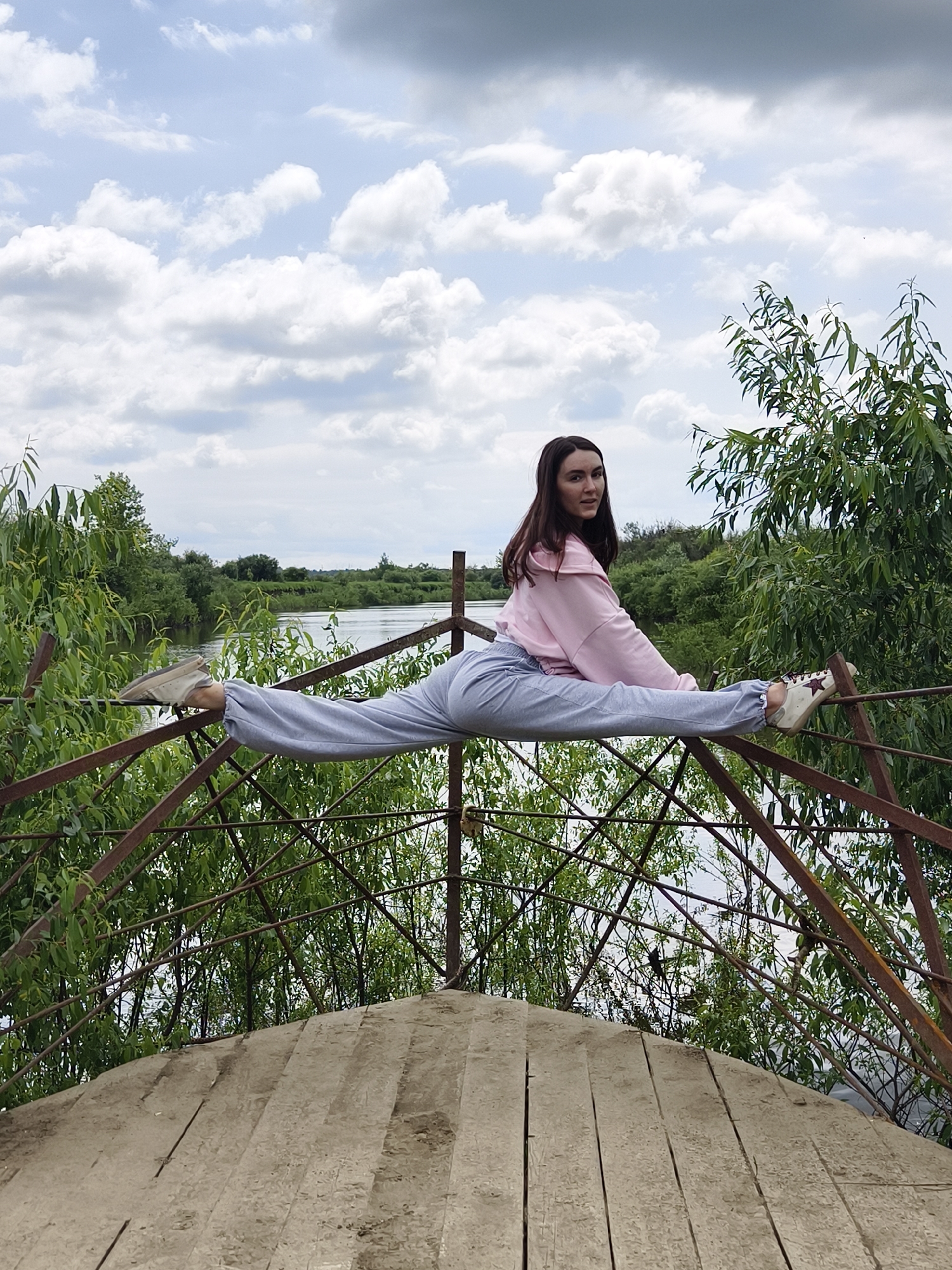 A young woman is performing a horizontal split while balancing on a rusty metal railing outdoors. She is wearing a light pink hooded sweatshirt and light grey sweatpants. Her legs are extended horizontally, with her feet resting on the railing. Her body is positioned to the left of the frame, and she is looking towards the camera with a slight smile. The railing is part of an outdoor structure, possibly a bridge or a viewing platform, which is supported by metal beams.
Behind the woman, a body of water is visible, bordered by lush green trees and foliage. The sky is partly cloudy, with a mix of white and gray clouds against a light blue background. The foreground features a wooden platform, made of wide planks. The scene appears to be in a natural, possibly park-like setting. The location is identified as the city of Онохино, Russia.