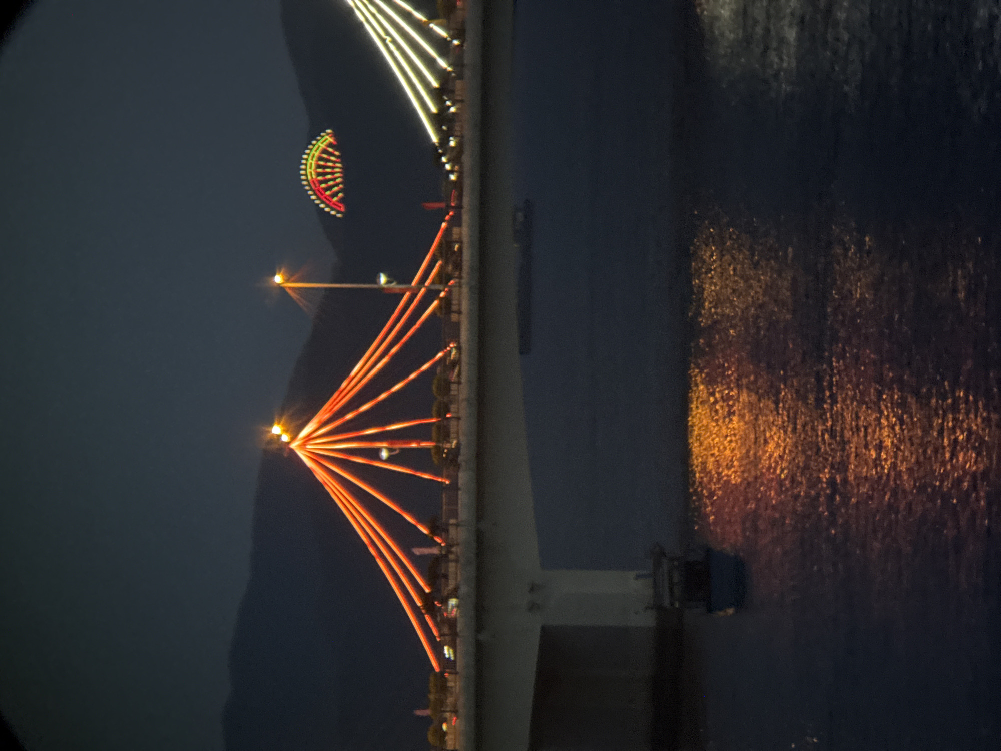 This is a nighttime photograph of a bridge illuminated by numerous lights. The bridge has a suspension design with cables radiating from tall towers. The cables and parts of the bridge structure are lit with orange and yellow lights, creating a striking visual against the dark sky. In the distance, another illuminated structure, possibly a Ferris wheel or another bridge feature, is visible with red and green lights. The bridge spans across a body of dark water, reflecting the colorful lights from above. A small boat is visible in the water below the bridge. The background shows a dark, mountainous silhouette. The scene is located in Nha Trang, Vietnam, and is likely the Tran Phu Bridge, which is a well-known landmark in the city, famous for its nighttime illumination.