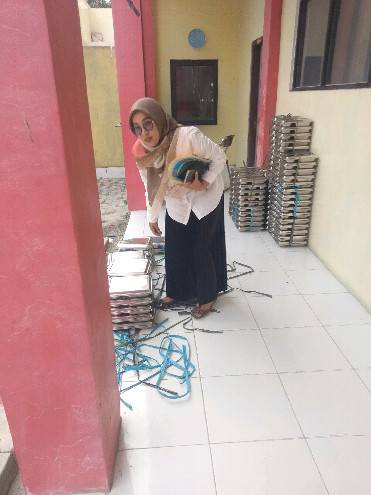 A woman wearing a hijab and glasses is bending over to pick up stacked trays outdoors. She is in the city of Suka Sari, Indonesia. The trays appear to be made of metal and are stacked neatly in rows. Some blue and green plastic straps are scattered on the white tiled floor around the trays. The woman is wearing a white long-sleeved shirt, a black skirt, and brown sandals. Behind her, there is a yellow wall with a dark window and a blue clock on it. To her left, a large pink pillar stands prominently. The scene suggests a setting related to food service or event preparation.