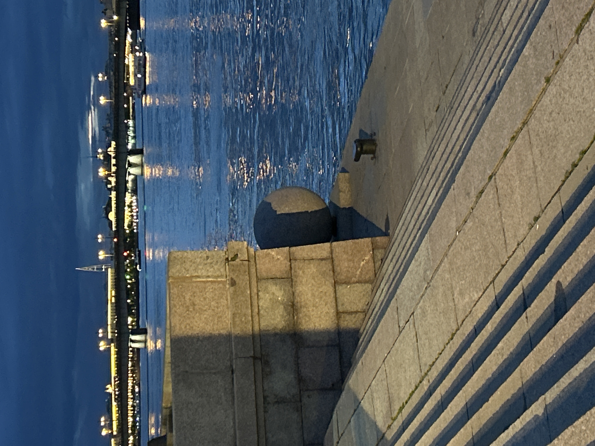 A wide, horizontal shot captures a waterfront scene at dusk, with a stone embankment in the foreground leading down to a body of water. On the left, a large, rough-hewn stone structure, possibly part of a retaining wall or monument, casts a long shadow across a set of stone steps. These steps, also made of large, irregular stone blocks, descend diagonally from the left to the right foreground, with distinct linear shadows falling across them, indicating a low light source from the left.
The stone embankment to the right of the steps is flat and grey, with a dark, rounded, spherical object positioned on it, partially obscuring the stone wall behind it. A short, dark metal bollard is visible on this embankment, close to the water's edge.
The water, likely a river or canal, is a deep blue, reflecting the lights from the cityscape across the water and the dim evening sky. Small boats with illuminated decks are visible on the water. In the background, a long bridge stretches across the water, lined with streetlights that cast a warm glow. Beyond the bridge, a city skyline is visible, featuring buildings and a distinctive modern spire, all silhouetted against the darkening sky. The sky itself is a deep, bruised blue, with some wispy clouds visible. The overall impression is one of a tranquil, urban waterfront at twilight.
