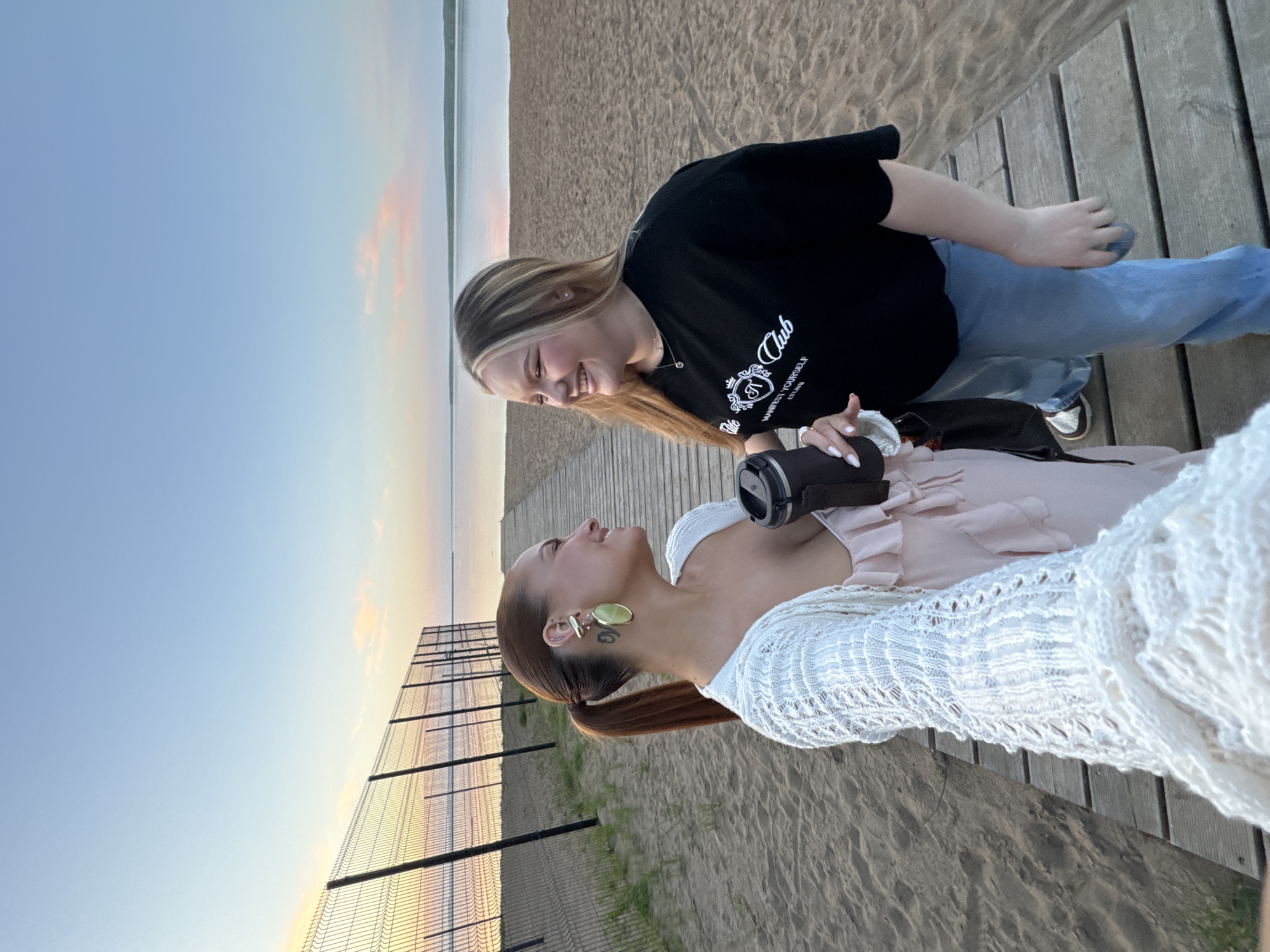 Two young women are on a wooden boardwalk near a beach. One woman wears a black t-shirt and jeans, and the other a pink top and white sweater. They are looking at each other and smiling. A fence is on the left and the beach and water are in the background. It is dusk. This image was taken in the city of Chaikovsky, Russia.