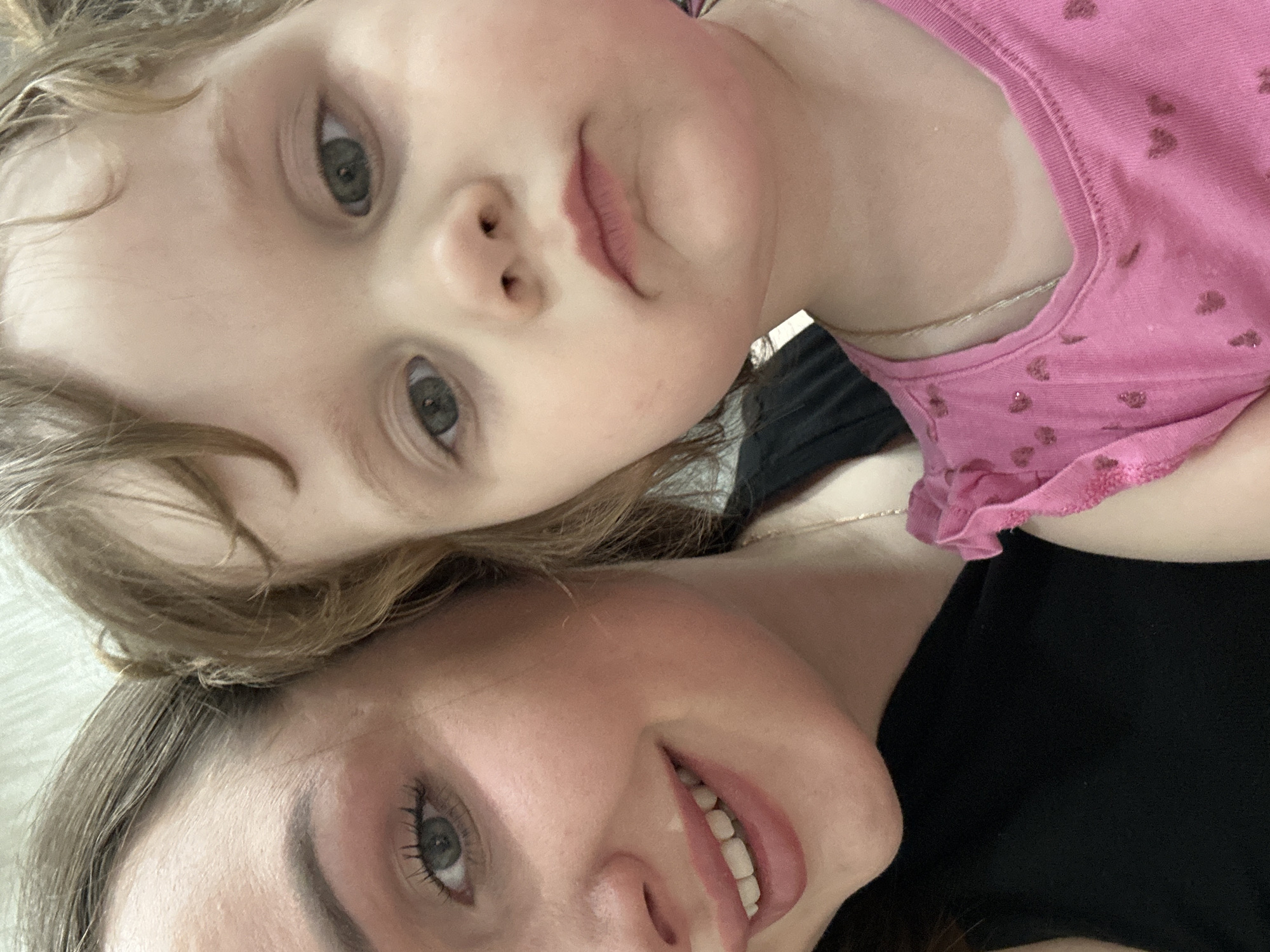 The image shows two people, likely a mother and child, posing for a selfie. The child, wearing a pink shirt with small heart patterns, is positioned above and slightly to the left of the woman. The woman is wearing a black shirt. Both are looking at the camera. The background is out of focus and appears to be a light-colored surface. There is no evidence of the location being in Chaikovsky, Russia.