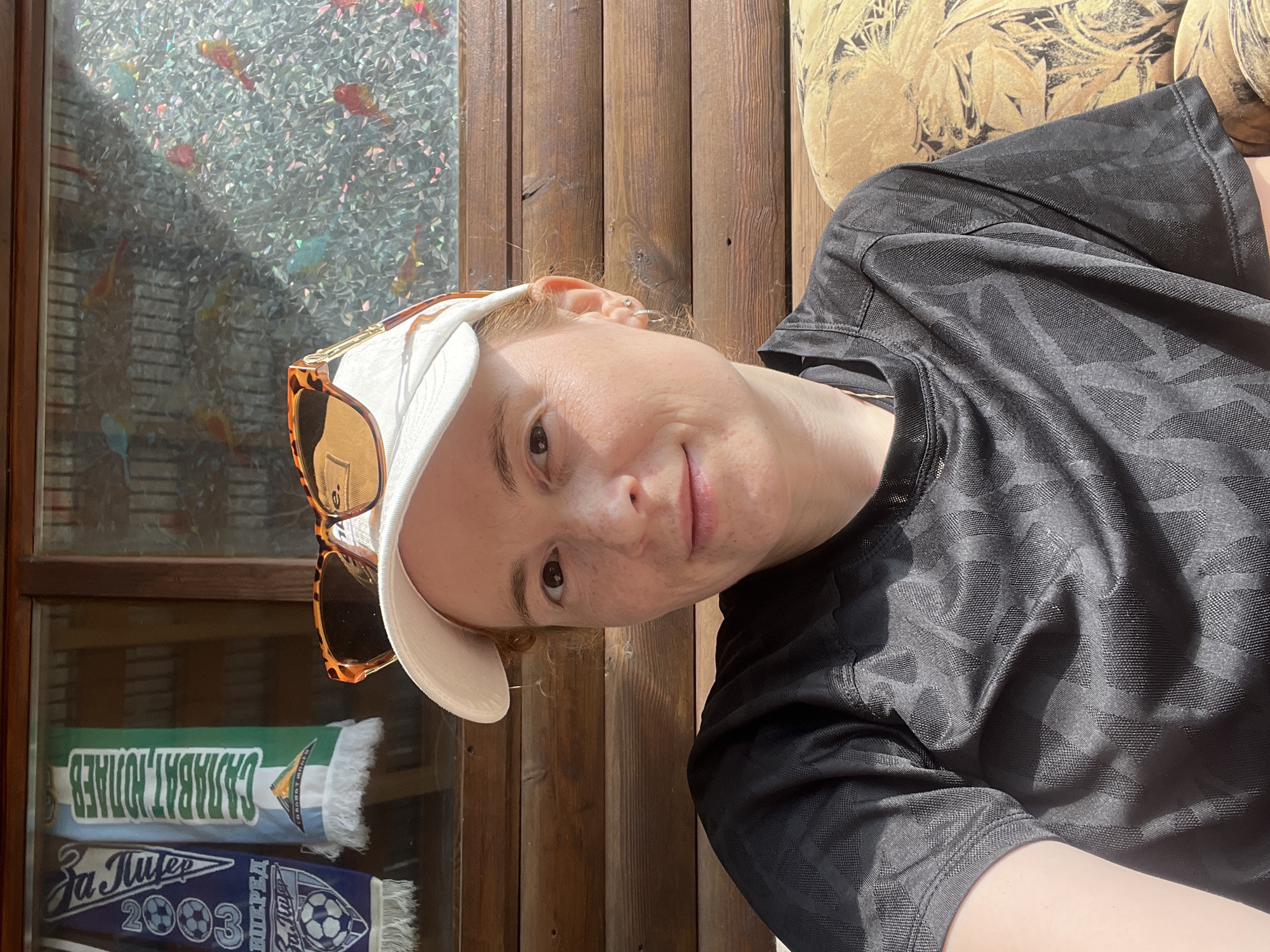 A medium shot depicts a person with freckled skin and reddish hair, wearing a white baseball cap and sunglasses perched on the cap. They are seated in front of a paneled wooden wall next to a window featuring patterned glass. Behind the window, two soccer scarves are visible, reading "САЛАВАТ ЮЛАЕВ" and "За Лидер", with the year "2003" and a soccer ball logo on the latter. The person is wearing a black patterned shirt and appears to be resting on a yellow patterned cushion. The location is likely Chaikovsky, Russia, based on the Cyrillic text on the scarves.