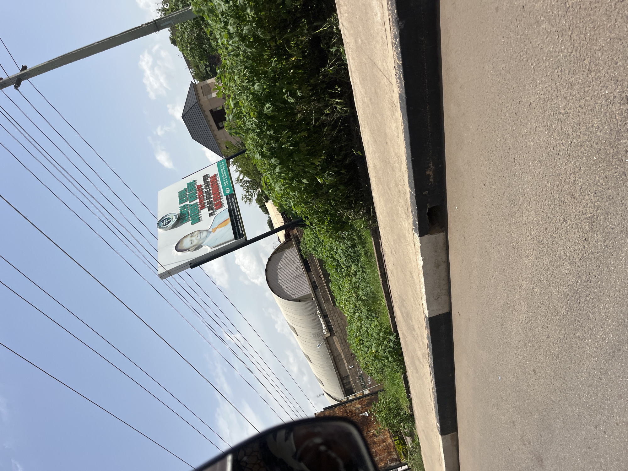 The media file shows a vertical view of a street scene in Gura Top, Nigeria. The dominant subjects are a billboard, lush green vegetation, power lines, and a street. The billboard, positioned on the left side of the frame, features text and an image of a person. The text on the billboard appears to be in green and red, with some parts obscured. It includes what seems to be a business name and contact information such as "08155859593" and "07032070721." The green vegetation is dense and overgrows along a wall bordering the street, suggesting a potentially humid climate. Power lines crisscross the sky, a common sight in many urban and semi-urban areas. A grey concrete curb separates the road from the vegetation. The road itself is paved and appears to be a standard asphalt surface. In the background, a building with a dark, angled roof is visible, partially obscured by the trees. Further back, a long, curved structure with a corrugated metal roof suggests an industrial or commercial building. The sky is partly cloudy, indicating it's daytime. The overall impression is a typical street view in a tropical or sub-tropical region, with signs of development and infrastructure. The angle of the shot, from a low perspective looking upwards, might suggest it was taken from a vehicle.