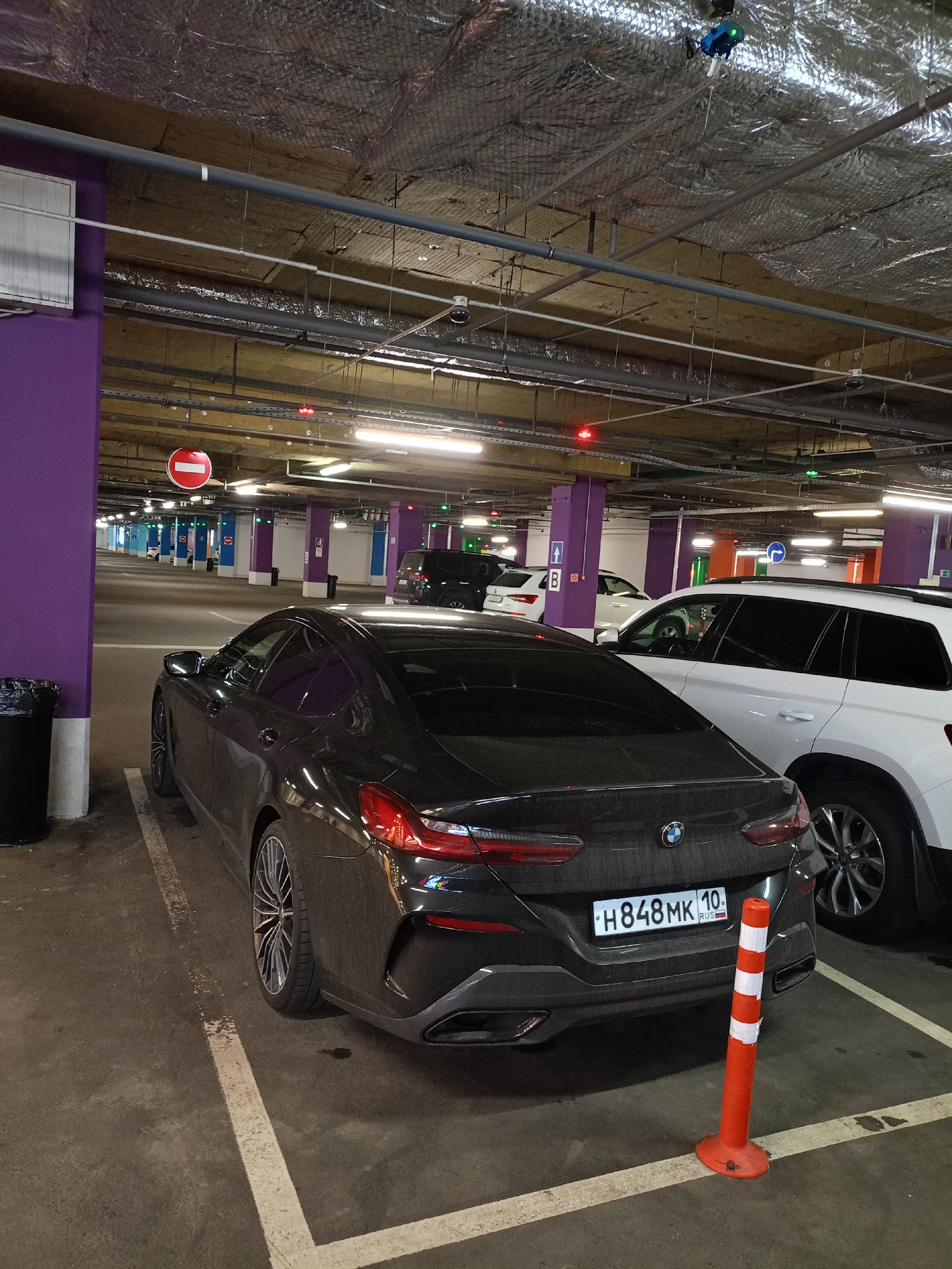 The image shows the interior of an underground parking garage. In the foreground, a dark gray BMW sedan is parked in a marked spot. The car's rear is facing the viewer, and its license plate reads "H848MK 10 RUS". To the right of the BMW, a white SUV is partially visible, parked in an adjacent spot. The parking garage features concrete floors, a suspended ceiling with exposed pipes and insulation, and fluorescent lighting. Purple painted pillars are present throughout the garage, with some displaying signage. An orange traffic cone with white reflective stripes is positioned in front of the BMW. In the background, other cars and parking spaces are visible, along with directional signs and illuminated indicators on the ceiling. A trash bin is located to the left of the BMW, next to a purple pillar.