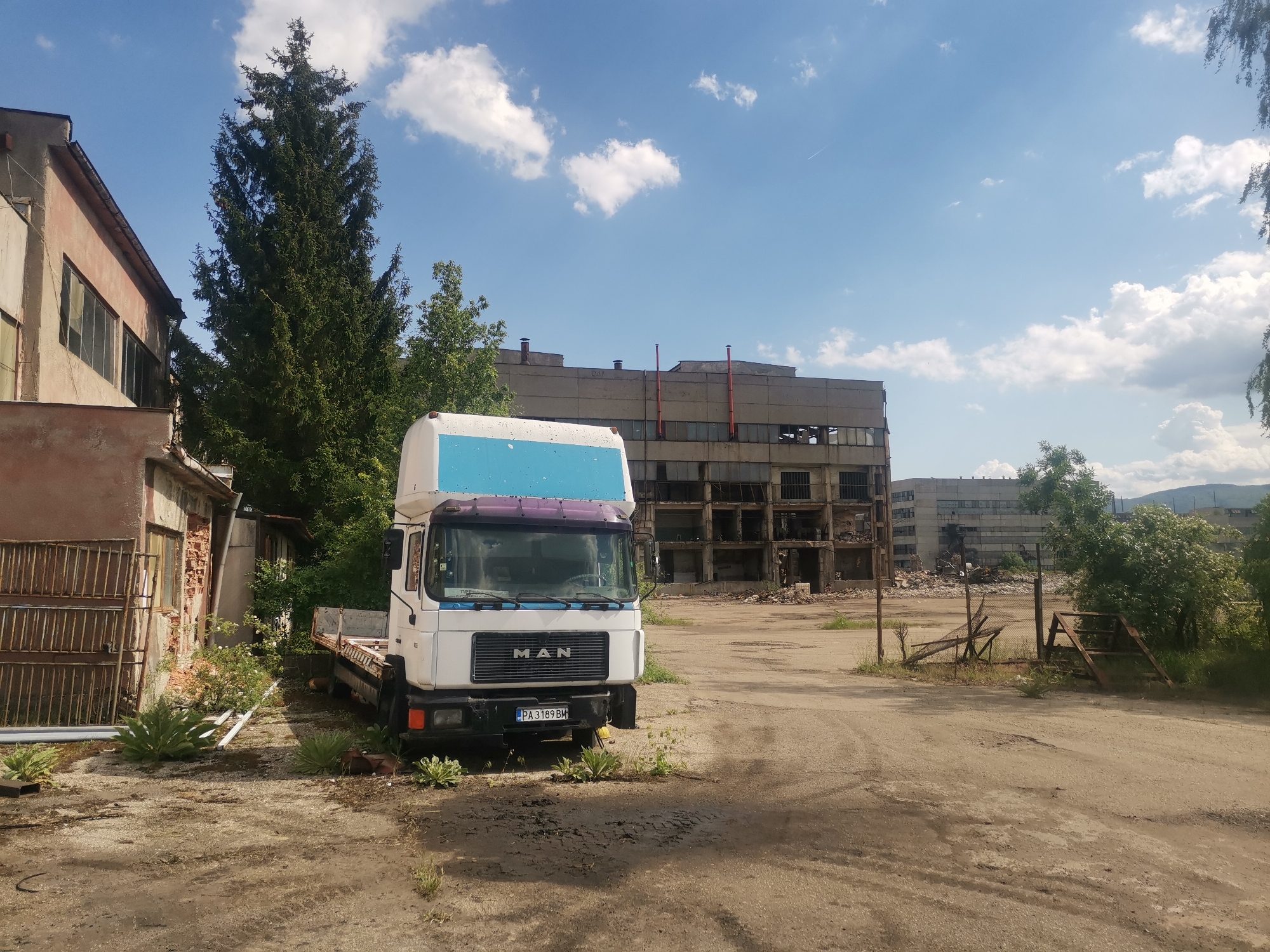The image depicts a weathered MAN truck parked on a dirt lot in front of a decaying industrial building. The building appears heavily damaged with missing windows and crumbling concrete. The truck is white with a blue and purple cab roof. The license plate reads "PA 3189 BM". Vegetation grows sparsely around the truck and along the edges of the lot. A smaller, older building stands adjacent to the truck on the left. The sky is partly cloudy. The location is Zhivkovo, Bulgaria.