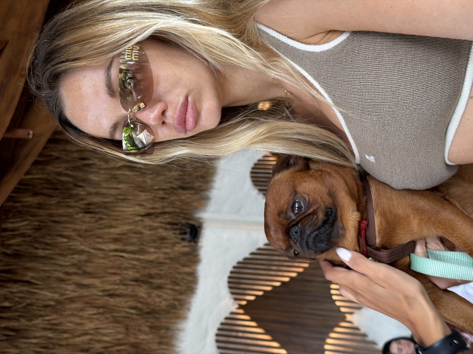 The image captures a blonde-haired woman holding a small, brown Brussels Griffon-type dog with a dark muzzle. The scene is located in Tibubeneng, Indonesia. The woman wears brown-tinted Miu Miu brand sunglasses and a ribbed, beige crop top with white trim. Her left hand, displaying white nail polish, rests on the dog's chest, which is fitted with a brown harness and a light blue leash. The dog looks forward and slightly left. A gold chain necklace is visible around the woman's neck. The background features a thatched roof section in the upper frame, and a decorative slatted wooden structure with a wavy pattern behind the subjects. A white fabric structure is partially visible further back, and a partially obscured individual is present in the lower-left background.