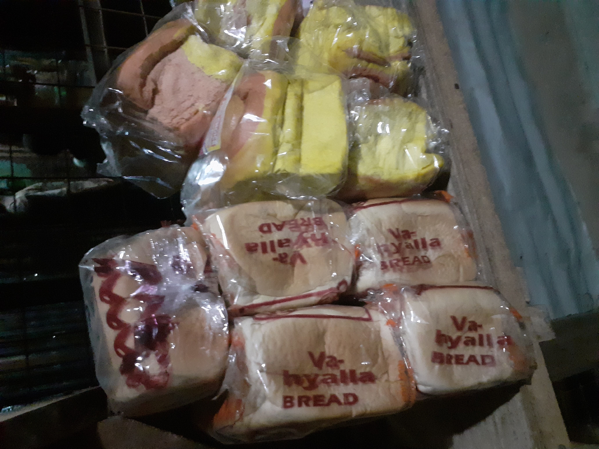 In Jalingo, Nigeria, a stack of packaged bread loaves is displayed on a wooden shelf. The loaves are wrapped in clear plastic, with some showing visible red markings or designs on top. The bread appears to be white and possibly yellow or light brown in color. The background is dark and somewhat blurred, suggesting an indoor setting or nighttime. The overall scene depicts a retail display of baked goods.