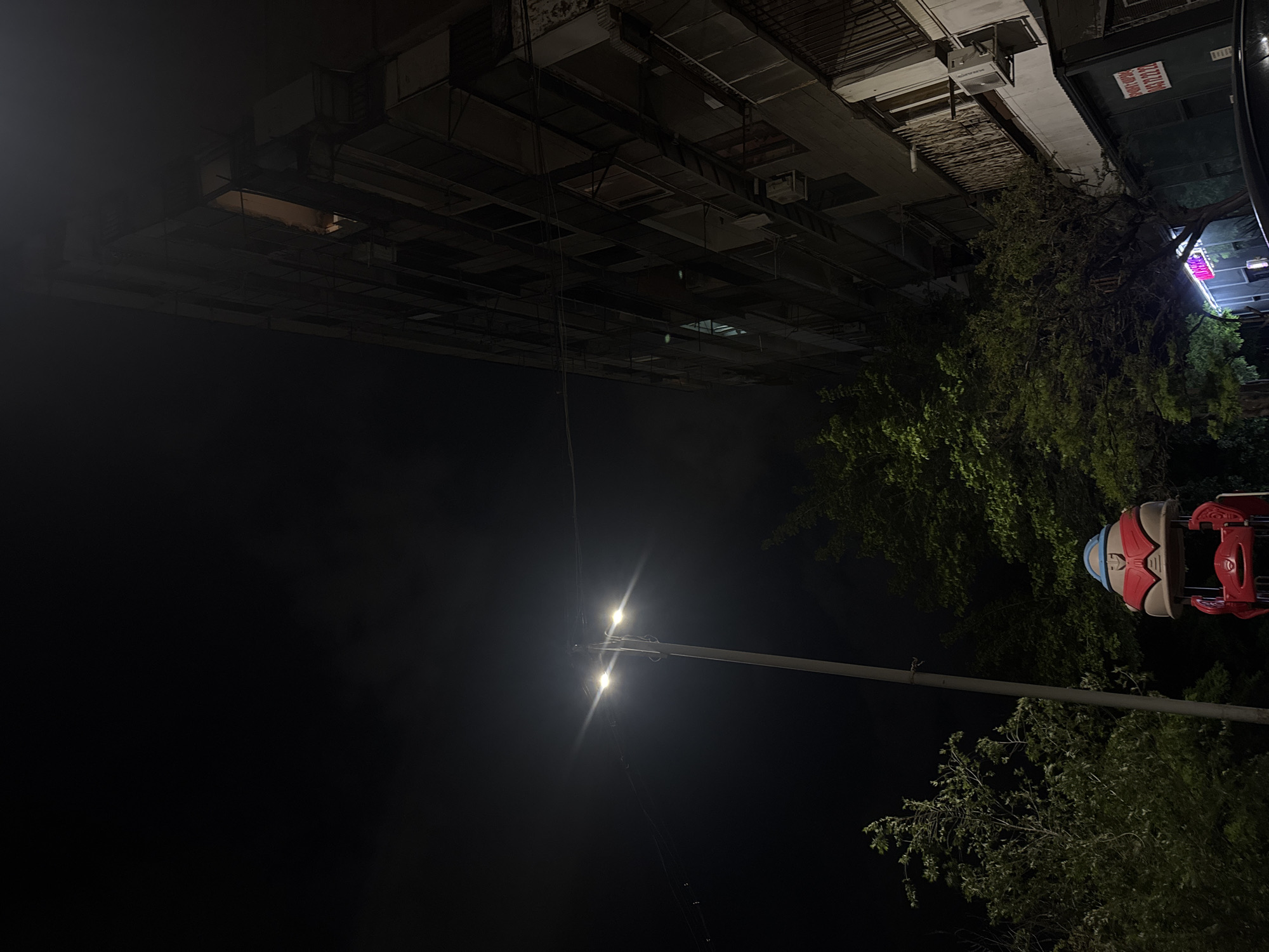 At night in Yerevan, Armenia, a low-angle view shows a multi-story building overhead. A street light shines brightly, and a red and blue ride is partly visible amidst trees on the right. A narrow, possibly vertical, sign with faint red text and white letters is visible at the edge of the building on the right. The sky is dark.