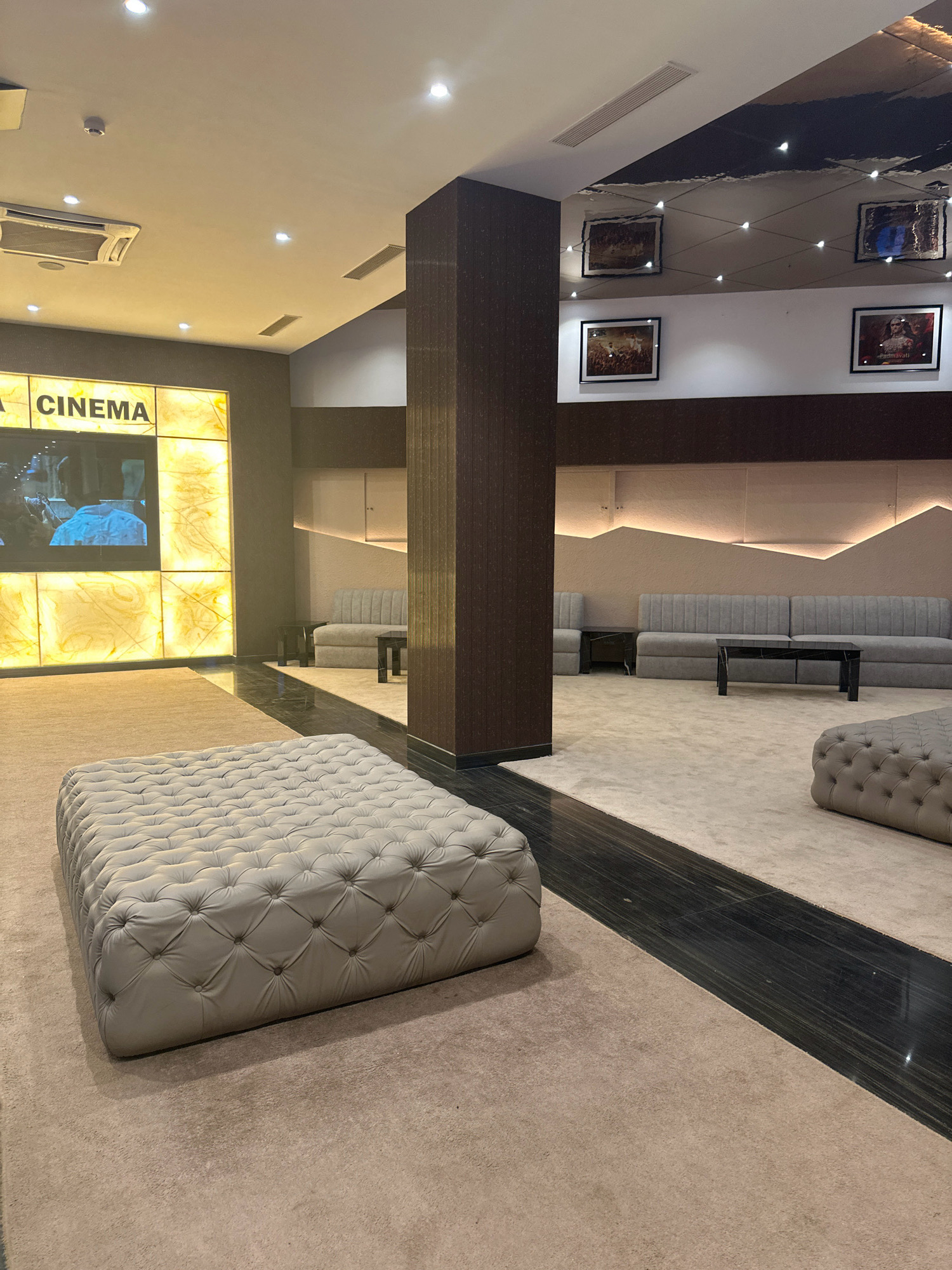The image shows the interior of a cinema lobby in Lalitpur, Nepal. In the foreground, a large, tufted ottoman upholstered in light gray leather sits on a beige carpet. Behind the ottoman, a dark, reflective strip runs across the floor. To the left, a large, backlit panel displays the word "CINEMA" in black lettering. The panel is illuminated by yellow light and features abstract, marbled designs. A portion of a screen showing a movie scene is visible within the panel. Further into the lobby, a central pillar with a dark, textured finish divides the space. Beyond the pillar, on the right side of the image, are two seating areas. One area features a long, tufted gray sofa with a dark, rectangular coffee table in front of it. A second, smaller tufted gray sofa is partially visible to the left of the larger one. The walls in this area are adorned with geometric lighting fixtures and framed artwork depicting historical or cultural scenes. The ceiling features recessed lighting and an air conditioning unit. The overall aesthetic is modern and luxurious.