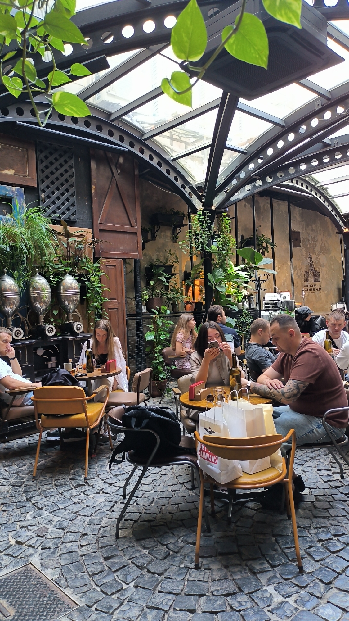 The image depicts a crowded outdoor seating area of a cafe, likely in Lviv, Ukraine, as suggested by the presence of Ukrainian text on a wall mural. The scene is characterized by a rustic, urban courtyard setting with a glass-paneled, arched roof structure. Cobblestone pavement covers the ground. Several patrons are seated at tables, engaged in dining and conversation.
In the foreground, a man with tattoos on his arm is seated at a table, facing right, wearing a brown t-shirt. A woman with a phone in her hand is seated at a table to his left, wearing a beige jacket. Another woman with blonde hair is seated at a table further left, wearing a white top. To her left, a man in a white t-shirt is seated.
The cafe's interior is adorned with numerous potted plants, creating a lush, green atmosphere. Two large, silver, vintage-looking coffee dispensers are visible on a shelf in the background. The walls are textured and appear to be made of aged plaster. A mural with Ukrainian text and an illustration of a building is visible on the right-hand wall. The overall ambiance is one of a lively, urban cafe with a bohemian or garden-like feel.