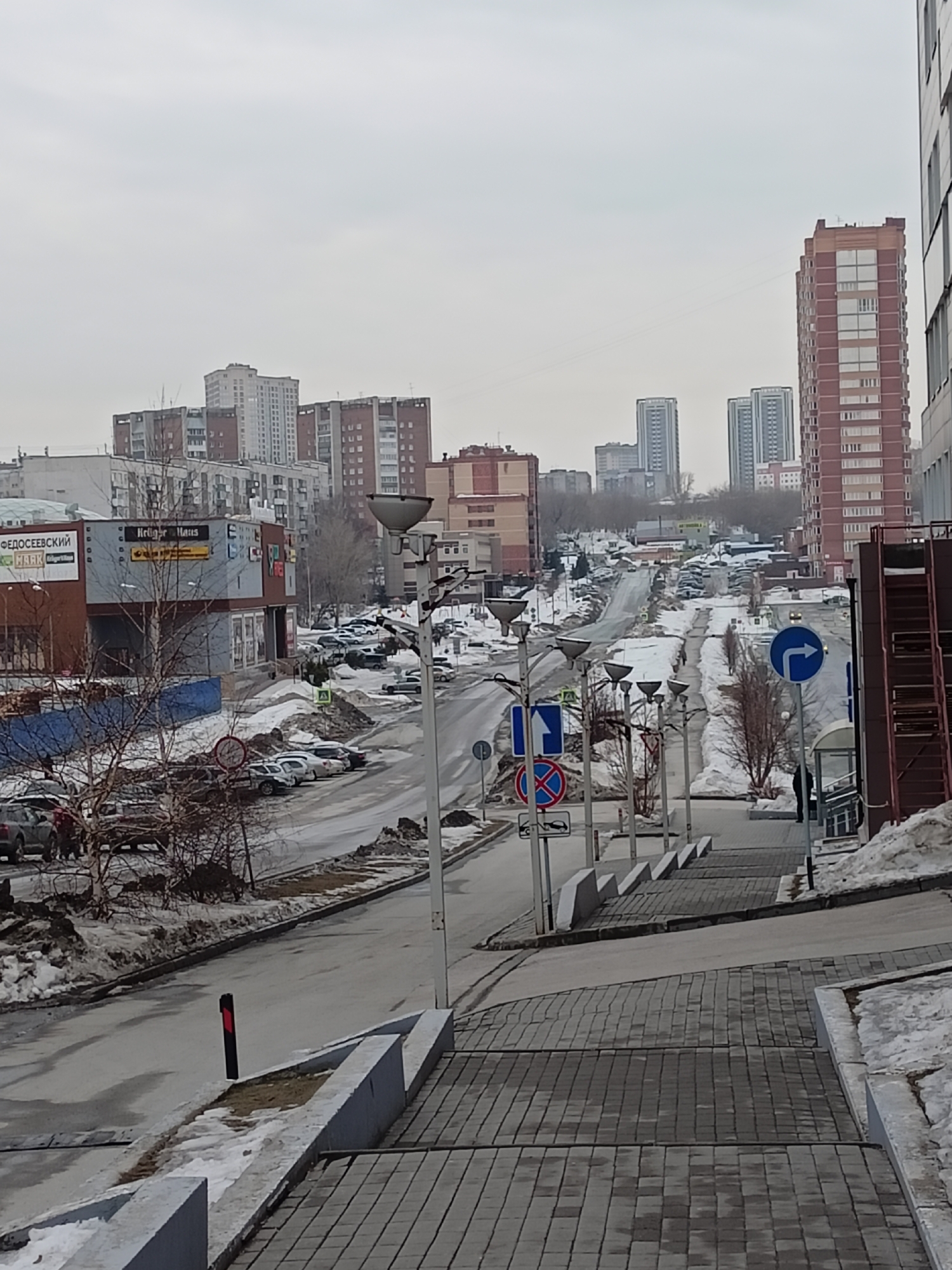 The image shows a cityscape in Novosibirsk, Russia on an overcast day. The view is from an elevated position looking down a paved pedestrian walkway with steps leading to a street. The walkway is lined with lampposts and street signs, including one indicating a right turn and another prohibiting parking for cars that do not meet certain requirements. Snow patches remain on the sides of the walkway and along the street.
On the left side of the street is a commercial building with signage, including "ФЕДОСЕЕВСКИЙ МЯСКО" and "Kreilinger Haus", adjacent to a parking area with multiple vehicles. The street ascends a slight incline, with parked cars lining both sides. In the background, multiple multi-story residential and commercial buildings can be seen against the overcast sky. There are a few bare trees interspersed within the urban environment. One person can be seen walking on the left.