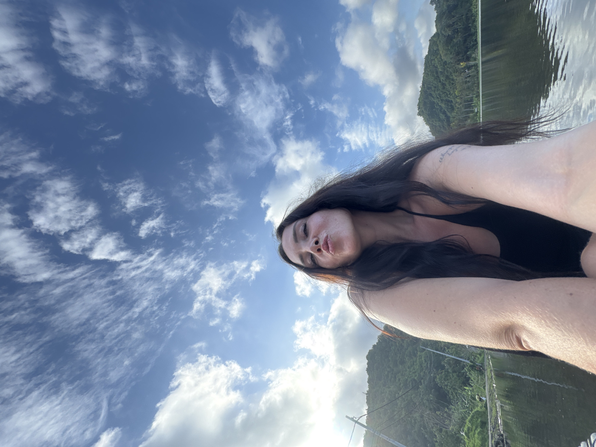 A woman with dark hair and a black top is taking a selfie in a body of water, with a lush green hillside and a bright blue sky with white clouds in the background. The sun is shining, casting a light on the scene, and there are some power lines visible on the left side of the image. The water reflects the sky and the surrounding landscape. The image appears to be taken in Manggis, Indonesia, given the tropical vegetation and atmosphere.