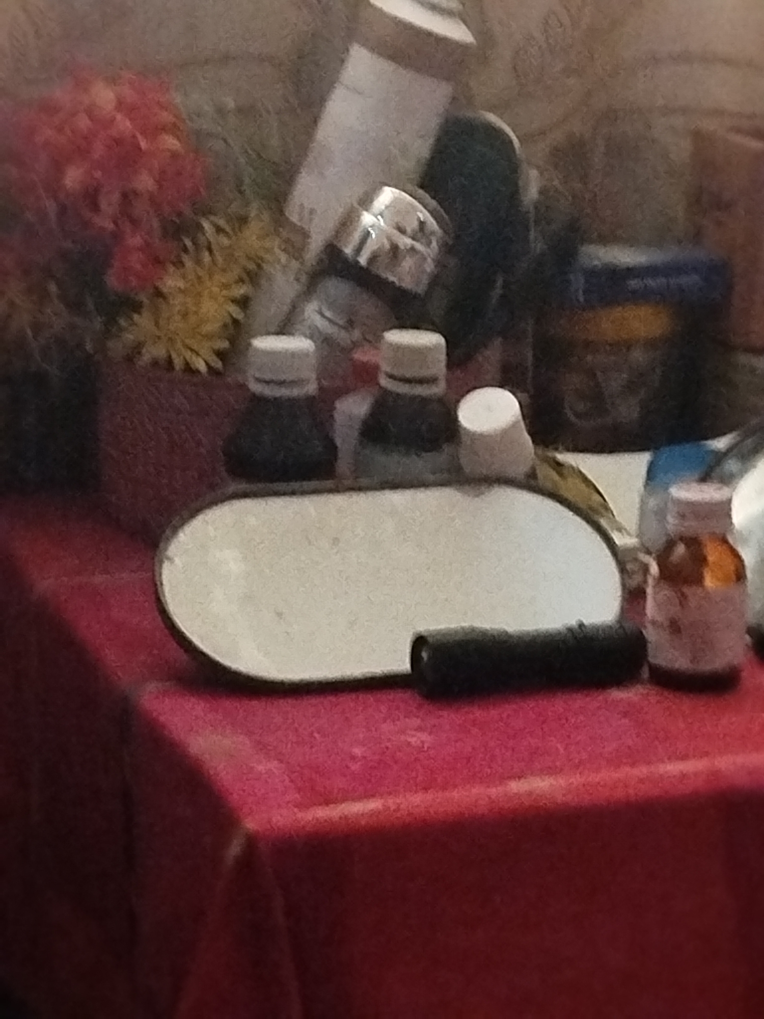 The image depicts a still life arrangement on a red tablecloth. Prominently featured is an oval-shaped mirror with a white surface, positioned horizontally. To the left of the mirror are two dark bottles with white caps. To the right of the mirror is a small, light brown bottle with a pinkish-white cap and a label. In front of the mirror and slightly to the right is a black cylindrical object, possibly a flashlight or a pen. In the background, there are various items including a white cylindrical object, a dark blue container with a yellow label, and a bouquet of artificial flowers in shades of red and yellow. The overall setting appears to be indoors, possibly on a dresser or table. There is no discernible information about the location within the city of Lassan, Pakistan, nor any indication of people or actions.