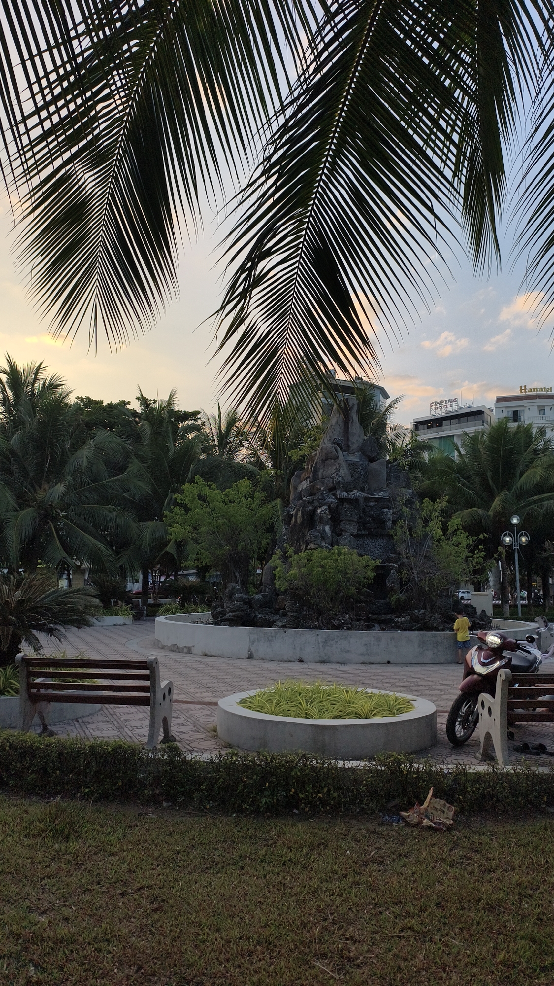 In an outdoor urban setting at dusk, a park scene unfolds with lush palm trees dominating the foreground, their fronds silhouetted against a soft, hazy sky. A paved walkway curves around a central circular planter filled with vibrant green foliage. Adjacent to the planter is a concrete bench, its weathered appearance suggesting it's well-used. Further along the path, a small, man-made rock formation resembling a waterfall stands prominently, surrounded by greenery. A maroon scooter is parked near another bench, and a lone individual in a yellow shirt stands by the rock formation, looking towards the park's center. In the background, modern buildings with signage, including "SPRING HOTEL" and "Hanatel," rise above the tropical foliage, indicating a commercial area adjacent to the park. The ground is covered with a mix of patchy grass and fallen leaves, with a small pile of debris near the foreground. The lighting suggests the end of the day, with a warm, diffused glow.
