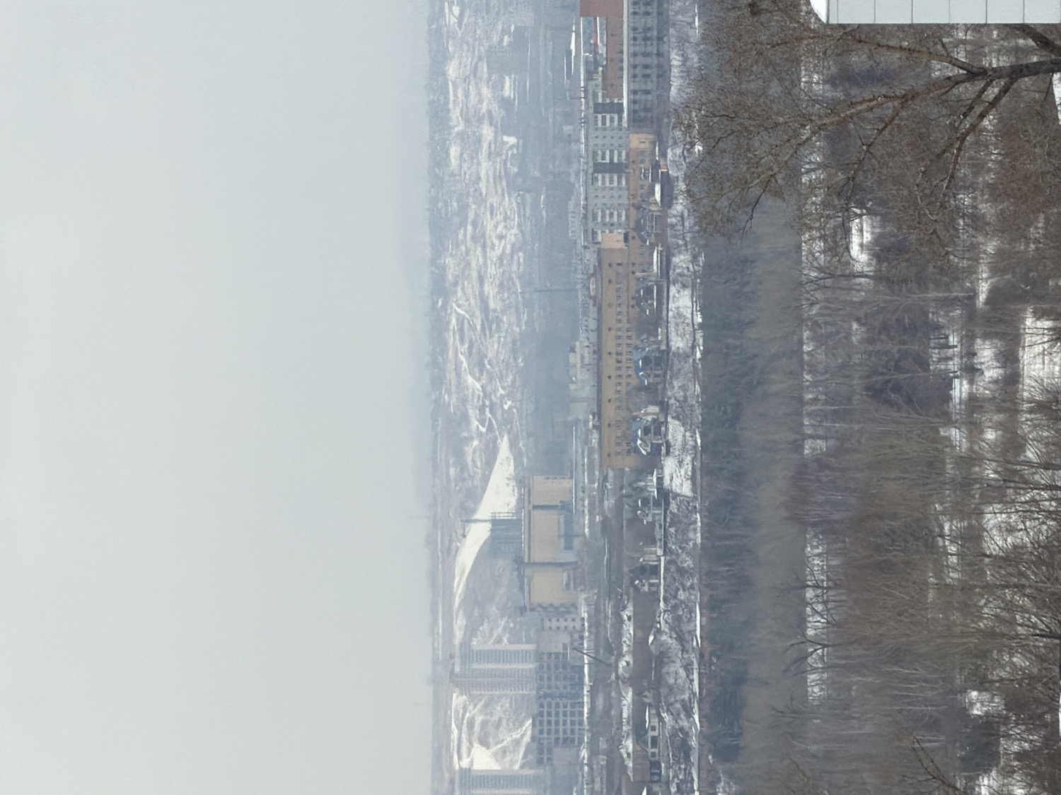 This media file presents a panoramic, high-angle view of an urban landscape alongside a wide river, under heavily overcast conditions. The image appears to be rotated, and if viewed with the large gray area at the top, it depicts a city in winter or early spring.
The upper portion of the scene is dominated by a vast, uniform expanse of light gray, indicating a heavily overcast sky or thick fog that reduces visibility. A wide river, likely partially frozen or with ice, cuts horizontally across the midground, its surface reflecting the dull sky.
On the left bank of the river, a dense urban area stretches into the distance. It comprises numerous multi-story buildings, including residential blocks of various heights and architectural styles, some light-colored, others a yellowish-brown. There are also smaller structures and what might be industrial or commercial buildings interspersed throughout the snow-covered ground.
The far bank of the river, on the right, features a wide strip of natural land covered by a dense array of bare trees, indicative of winter. Beyond these trees, the landscape appears snow-covered and gradually fades into the pervasive atmospheric haze. In the extreme upper right corner, a few bare branches from a closer tree are visible, suggesting the photo was taken from an elevated position, possibly a building or balcony, overlooking the scene.
The weather is cold and gray, with snow on the ground and bare trees, confirming a winter season. The heavy overcast sky and widespread haze lend a muted and somber atmosphere to the entire cityscape. No specific human activity or visible text can be discerned. The scene is characteristic of a large Russian city like Krasnoyarsk, situated on a major river during its cold season.
