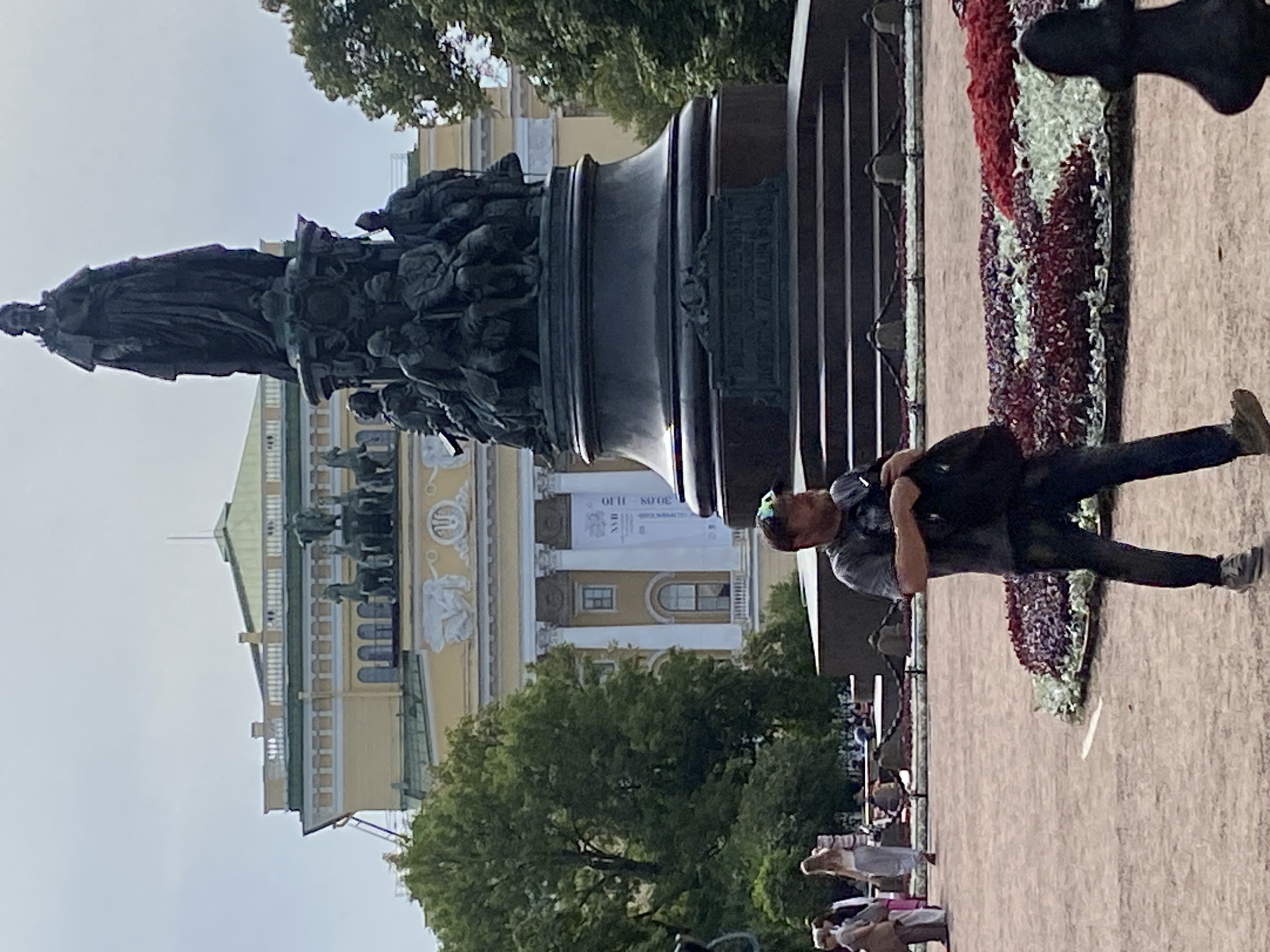 The image captures a portion of the monument to Catherine the Great in Palace Square, Saint Petersburg, Russia. In the foreground, a man is walking towards the right side of the frame, carrying a dark bag. He is wearing a grey t-shirt, dark pants, and sneakers. Behind him, a flowerbed with red and white flowers is visible. To the left of the man, two women are walking away from the camera. In the background, the monument to Catherine the Great stands tall, with its bronze figures and the statue of the empress. Behind the monument, a yellow building with classical architectural features, likely the General Staff Building, is partially visible. The sky is overcast. The overall scene depicts a public square with a prominent historical monument and people engaged in everyday activities.