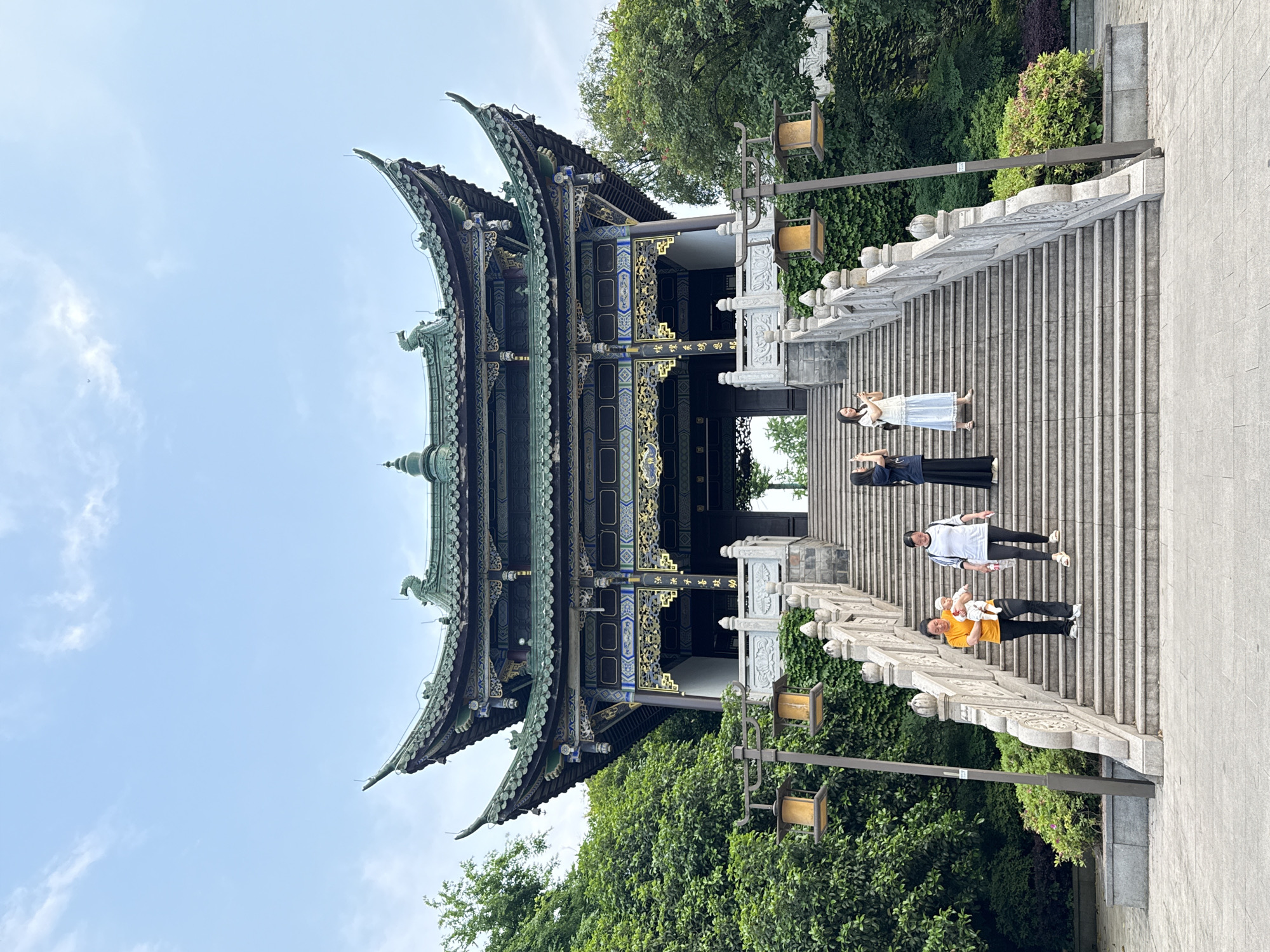 The image shows a multi-tiered traditional Chinese building, likely a temple or pavilion, in what appears to be a park-like setting in Liangjiang, China. The structure features ornate roofs with upward-curving eaves, detailed decorative elements, and a dark color scheme with gold accents. It is situated at the top of a long flight of wide, stone steps with carved stone railings.
Four people are ascending the stairs. The person closest to the camera is wearing an orange shirt and black pants and appears to be holding a baby. Above them, another person is wearing a white shirt with black stripes and dark pants. Further up the stairs, two women are standing and posing. One is wearing a white dress and the other a dark-colored top and pants.
The scene is surrounded by lush greenery, including trees and climbing vines on the railings. Lamp posts line the staircase. The sky is light blue.