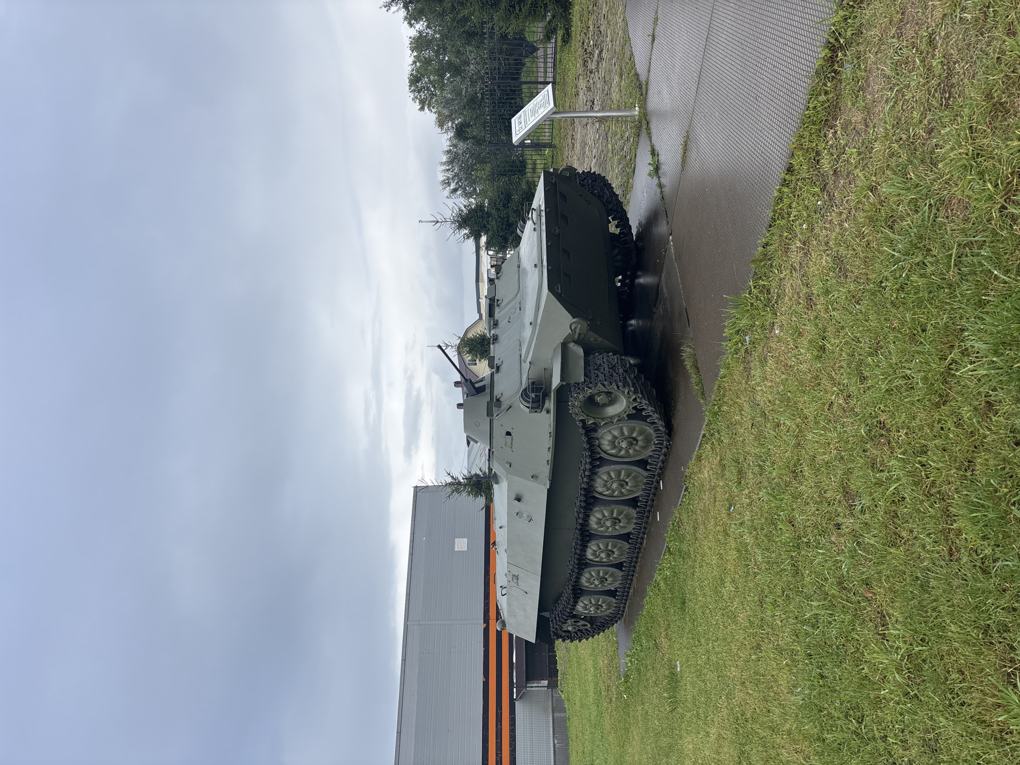 The image shows a military vehicle, a tracked armored personnel carrier, painted olive green, positioned on a paved surface with grass in the foreground and background. The vehicle has a turret with a machine gun mounted on top. Behind the vehicle is a large, rectangular gray building with an orange and black striped band running horizontally across its lower section. Several evergreen trees are visible behind the building and to the right of the vehicle. A black metal fence is also present in the background, with a sign on a silver pole in front of it. The sky is overcast and gray. The location is identified as the Luga, Russia city limits.