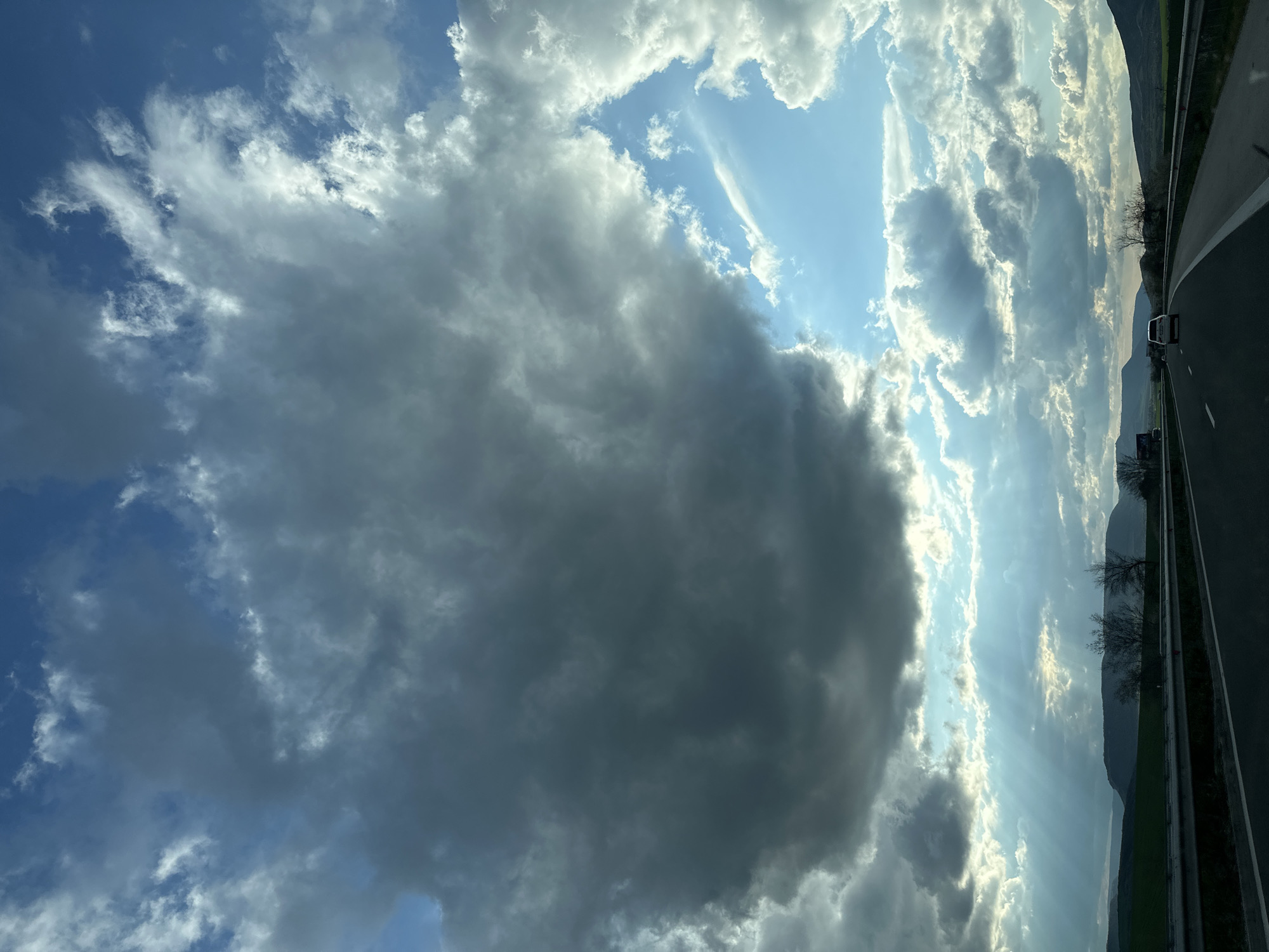 The image is a landscape oriented shot taken from a moving vehicle on a highway, presumed to be near Mlada Gvardia, Bulgaria. The left side of the image is dominated by a large, dark cumulonimbus cloud formation filling a substantial portion of the sky. The sky behind and around the cloud displays patches of blue interspersed with various other types of clouds. Sunlight appears to be streaming through some of the cloud formations, creating a visible light ray effect. The right side of the image depicts a highway with a guardrail separating the road from a green field and distant mountains. A single vehicle is visible on the highway, moving in the same direction as the camera. Bare trees are visible along the road.