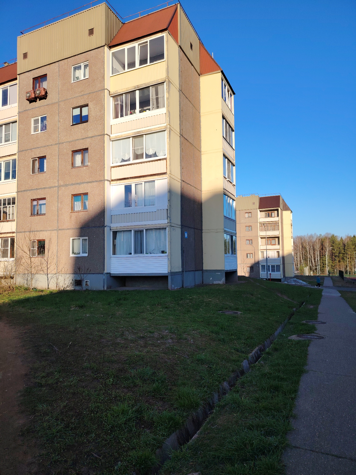 The image captures an outdoor scene in Марковский, Russia, featuring residential buildings and a surrounding environment under a clear blue sky.
In the foreground and mid-ground, two multi-story apartment buildings stand. The closest building, on the left, is constructed with large, gray concrete panels and features light yellow painted sections on its facade and enclosed balconies. It has numerous windows of varying sizes, some with white frames and others with brown frames. Two air conditioner units are mounted on one side of the building. The upper section of the building has a tan, corrugated metal structure, possibly housing a technical floor or attic, topped with a reddish-brown roof. On its far side, a blue sign with the number "10" is visible. The second, similar-looking building is further down the paved pathway on the right, sharing the same architectural style and reddish-brown roof.
The ground in front of the buildings and along the pathway is covered in green grass, appearing somewhat sparse in areas. A concrete or asphalt walkway extends into the distance, bordered by a concrete drainage ditch on the grass side. Several round manhole covers are visible on the walkway. In the mid-ground, a lone person dressed in a light blue jacket walks away from the viewer on this pathway. Bare trees or bushes are scattered near the base of the closest building, and a dense forest with leafless trees forms the horizon in the background.
The long shadows cast by the buildings suggest either early morning or late afternoon light, indicating a sunny day. The overall atmosphere appears calm and residential.