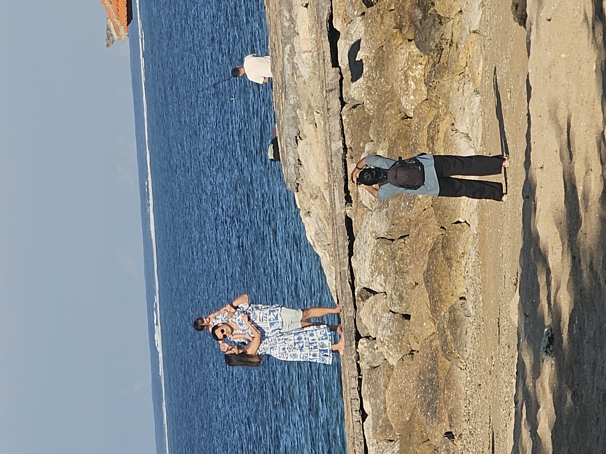 The image depicts a beach scene at what appears to be a resort or a public area in Denpasar, Indonesia, on a clear, sunny day. In the foreground, a person wearing a blue shirt and black pants with a backpack is photographing a family. The family, positioned on a rocky breakwater, consists of a man holding a child on his shoulders, and a woman standing beside them. The family is wearing matching blue and white patterned outfits. In the background, a person is fishing from the breakwater, and the ocean stretches to the horizon with a faint white line indicating a distant reef or breaking waves. A small portion of a building with an orange roof is visible on the right side of the frame. The ground is covered with sand and large rocks.