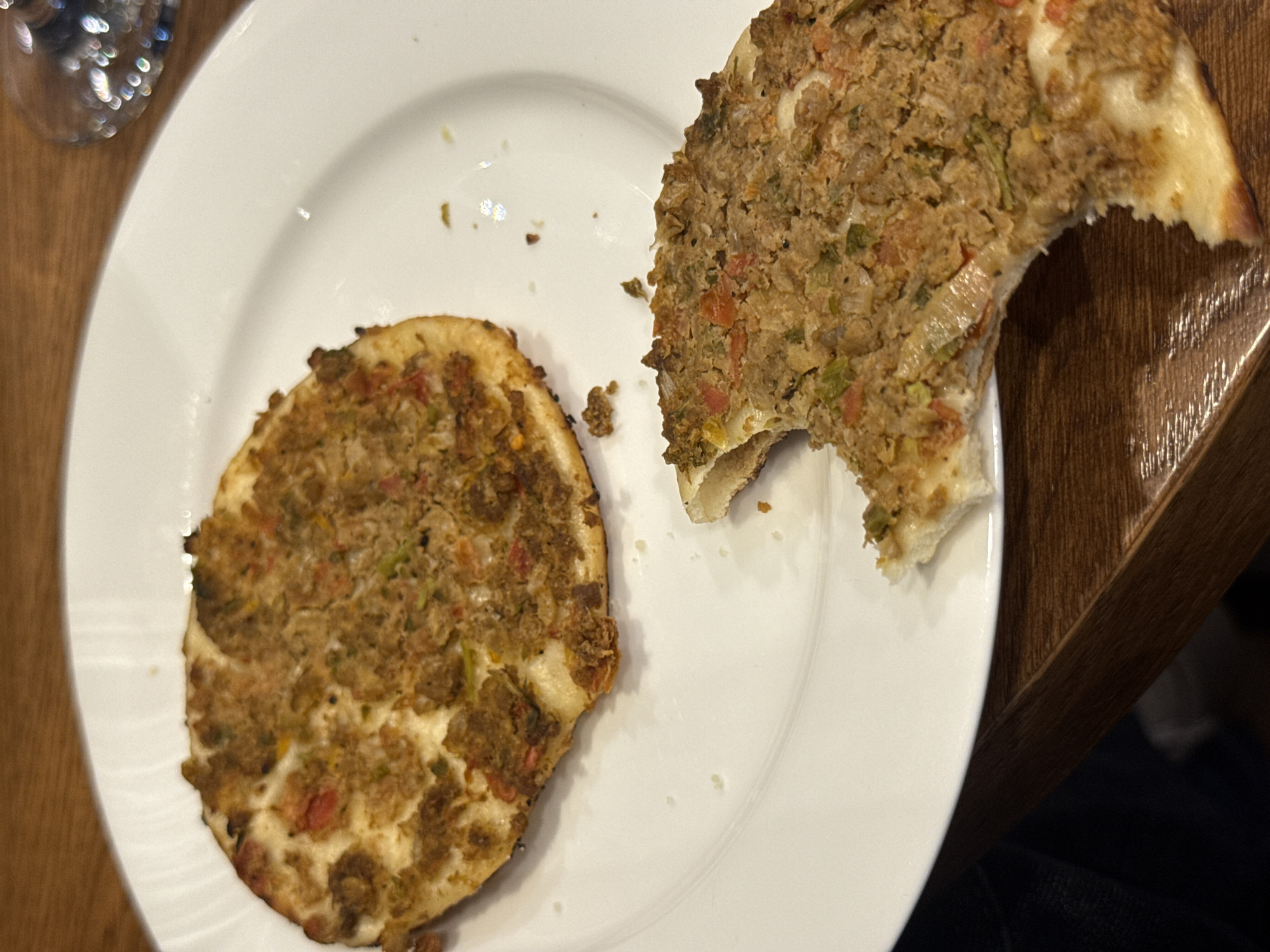 This overhead shot displays a white ceramic plate holding two pieces of a savory flatbread dish, likely taken in an indoor setting in Yerevan, Armenia. The dish strongly resembles Lahmajoun or Sfiha, characterized by a thin dough base topped with a cooked mixture of ground meat, finely diced tomatoes, and green vegetables, possibly onions or peppers. The topping exhibits a crumbly, slightly browned texture, while the underlying dough is pale with some crisp, golden edges.
One piece of the dish is a complete, circular portion, while the other is a larger, crescent-shaped piece from which a substantial bite has been taken, indicating the meal is in progress or has recently been consumed. A few small crumbs are scattered on the clean white surface of the plate.
In the upper-left corner, a blurred clear glass, possibly a wine glass, with shimmering light reflections is partially visible. The plate rests on a warm-toned wooden table with a distinct grain pattern, visible on both sides of the plate. A patch of dark fabric, likely clothing, is just visible in the bottom right corner, suggesting a person is seated at the table. The overall lighting suggests an indoor setting, possibly during an evening meal. There is no visible text in the image.