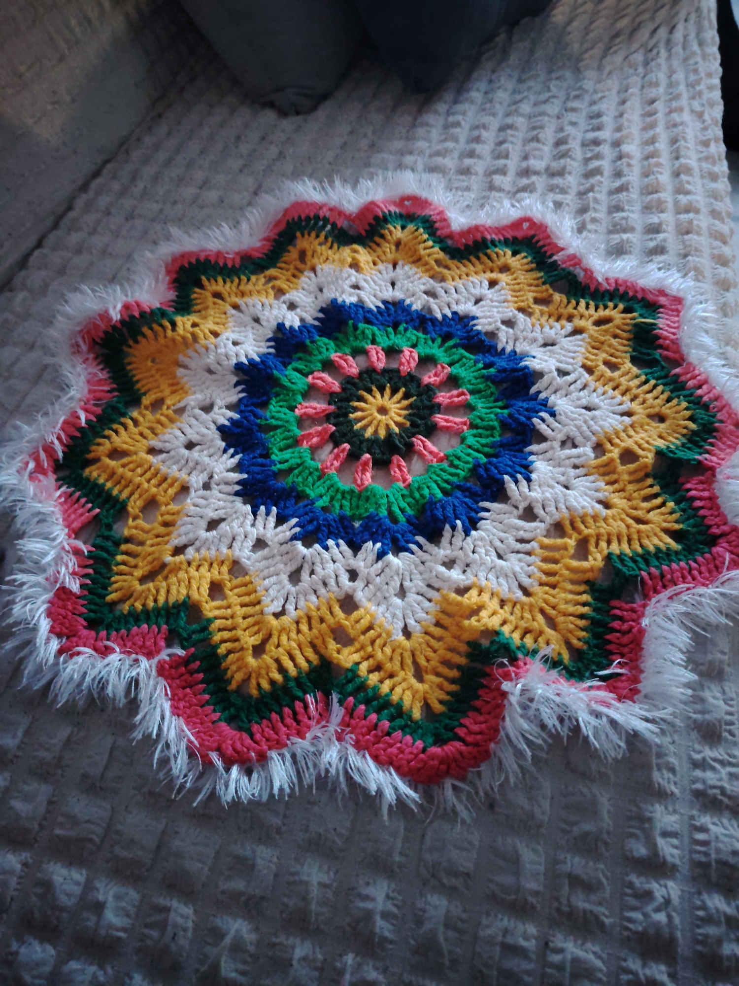 The image displays a close-up, elevated view of a brightly colored, circular crocheted item, likely a placemat or doily, resting on a textured, light-colored blanket or bedspread. The crochet piece exhibits a complex, multi-layered mandala-like pattern with a central star-shaped motif.
The colors of the crochet work are vibrant and varied, including a dark center, surrounded by rings of light pink, green, blue, white, yellow, and then alternating green, yellow, and white segments towards the outer edge. The outermost ring is predominantly a solid bright pink with a scalloped edge. A unique feature is the fluffy, white, textured yarn incorporated as a fringe along the entire perimeter of the pink edge, giving the item a soft, decorative border. The intricate crochet stitches are clearly visible, showcasing the handmade nature of the item.
The setting appears to be indoors, possibly on a bed or a sofa, suggested by the soft, patterned blanket beneath the crochet piece and the blurry, dark grey object (possibly a cushion or part of furniture) in the upper background. The lighting suggests ambient indoor light, possibly from a window, creating some subtle shadows on the blanket. There are no people present, and no specific activity is depicted; the image is a still life focused on the handcrafted object. No text is visible in the image. The location context for this media file is Сусады-Эбалак, Russia.