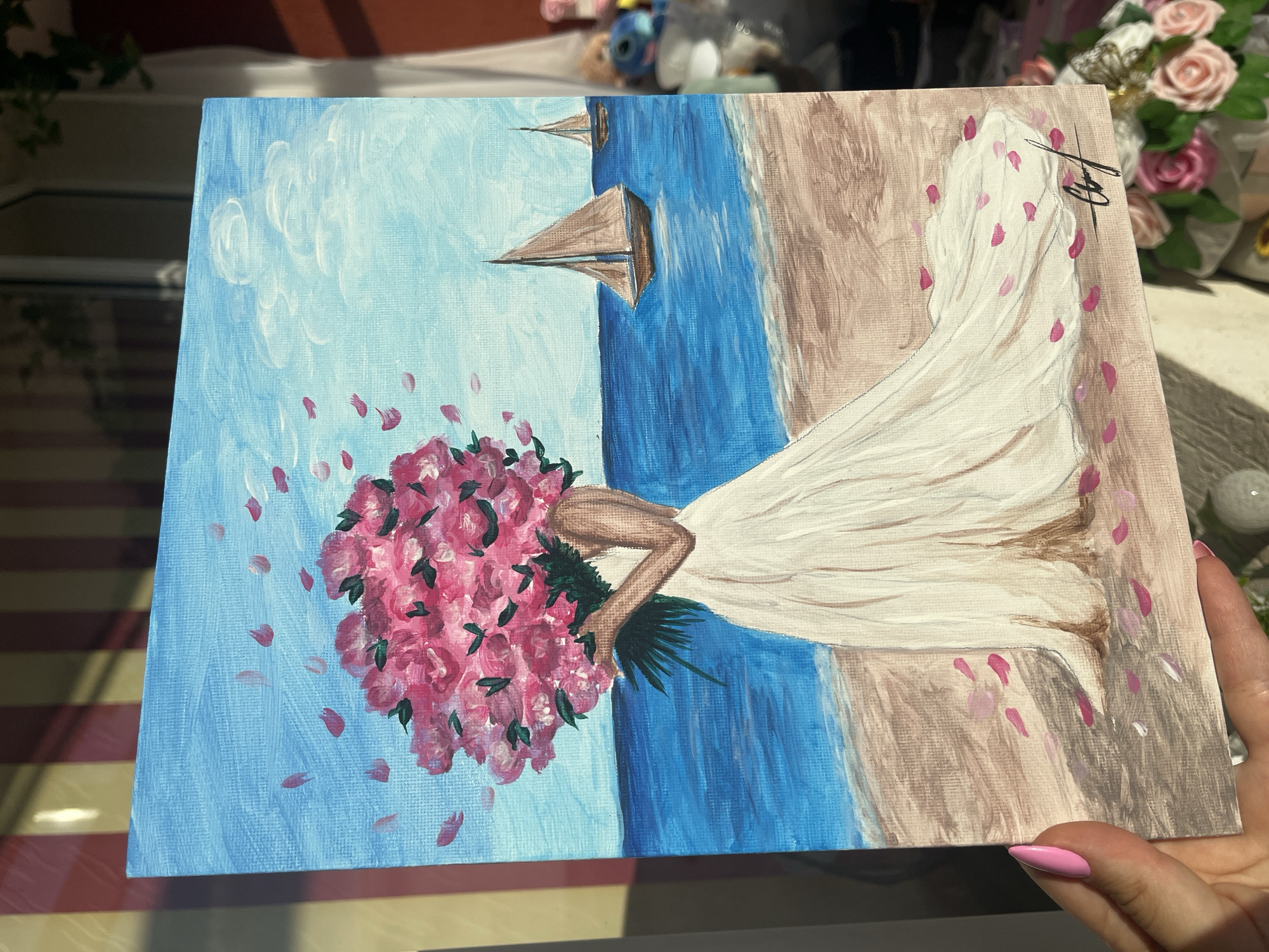 An acrylic painting on canvas depicts a scene with a woman in a white dress holding a bouquet of pink flowers on a beach. The background consists of a blue sky with white clouds, a blue sea with sailboats, and a tan-colored sandy beach. Pink flower petals are scattered around the woman and on the sand. The painting is signed in the upper right corner. The location in the city of Veliko Tarnovo, Bulgaria, is not discernible from the painting.