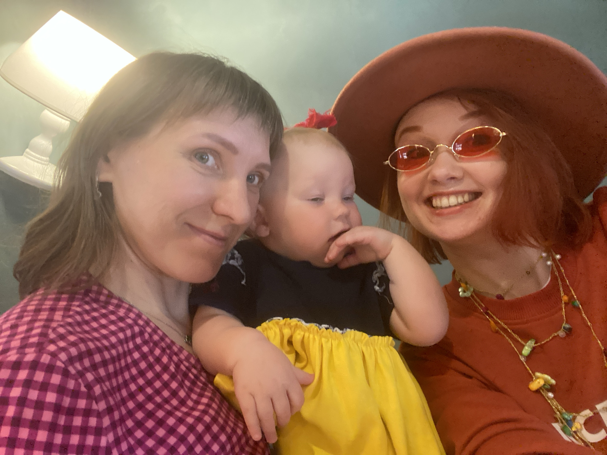 Three individuals are captured in a selfie-style photograph. On the left is a woman with short brown hair and light eyes, wearing a pink and maroon checkered top. In the center is a baby in a Snow White-themed outfit, consisting of a black top and a yellow skirt, with a red bow in their hair. The individual on the right is another woman with red hair, wearing a brown hat and orange-tinted sunglasses, along with a rust-colored sweater and layered necklaces. The background is indistinct, though a lamp is visible in the upper left corner. The image suggests an indoor setting. There is no indication in the image it was taken in Yekaterinburg, Russia.