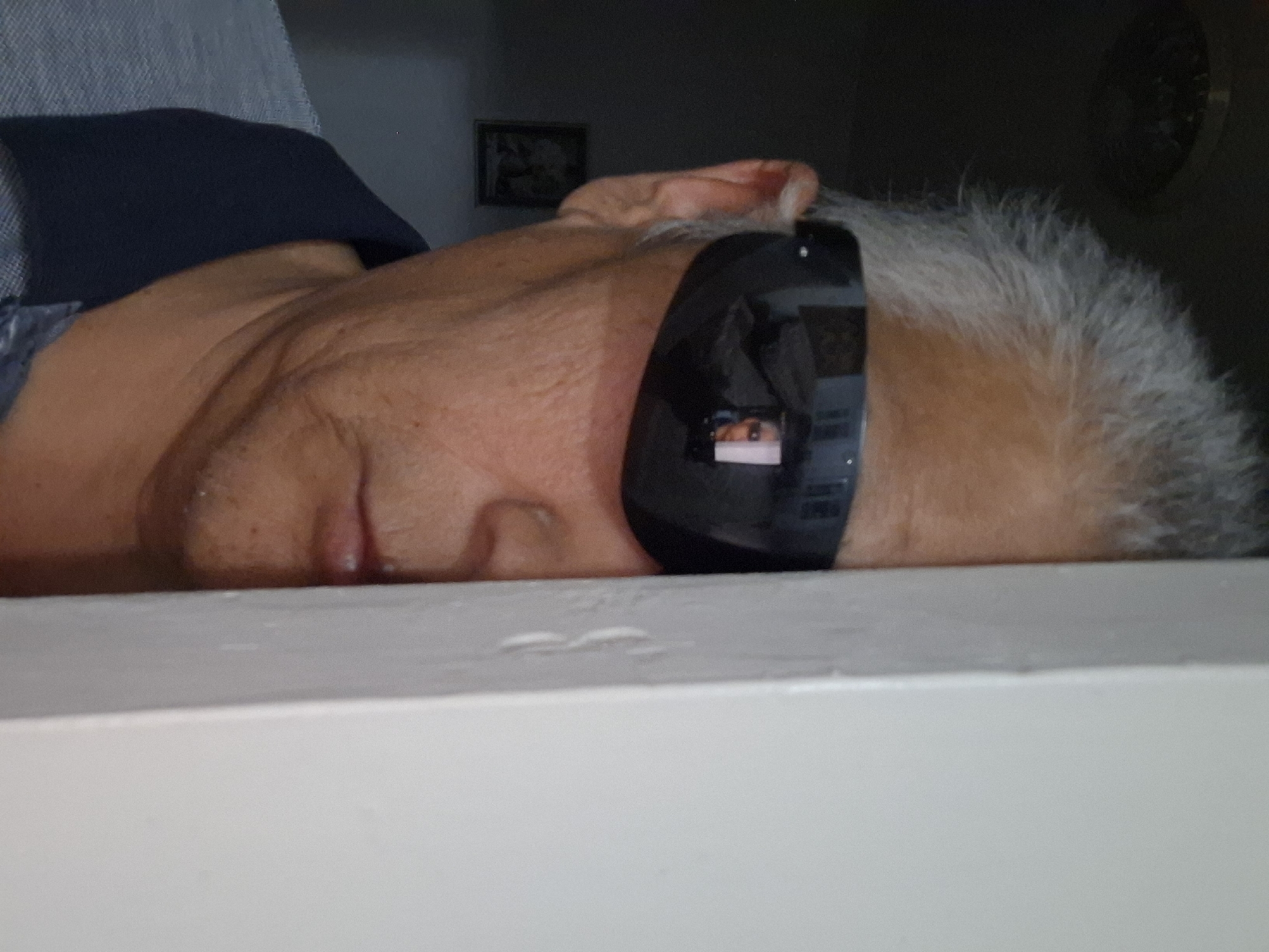 A close-up, portrait-style image shows a middle-aged East Asian man from the shoulders up, partially obscured by a light-colored vertical surface on the right side of the frame. The man has short, gray hair and is wearing dark sunglasses that reflect a room with furniture and a person. He is looking towards the camera and has his right ear and cheek visible. He is wearing a dark collared shirt. The background to the left of the man is dark, with a faint ceiling fixture visible at the top and a framed picture on the wall below it. The image quality is moderate, with some graininess. The setting does not provide any specific indicators of its location within the city of Almaty, Kazakhstan.