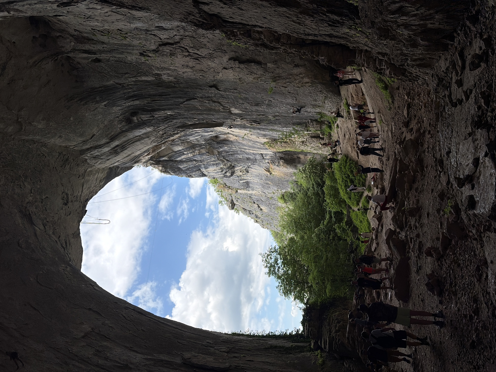 The image depicts a large cave entrance in Unknown, Unknown, framing a view of the sky with clouds. The cave walls are textured and rocky, and a group of people are visible along the cave's edge, some standing while others are climbing. There is a rope visible near the cave entrance, suggesting climbing activity. The environment outside the cave includes green trees and vegetation.