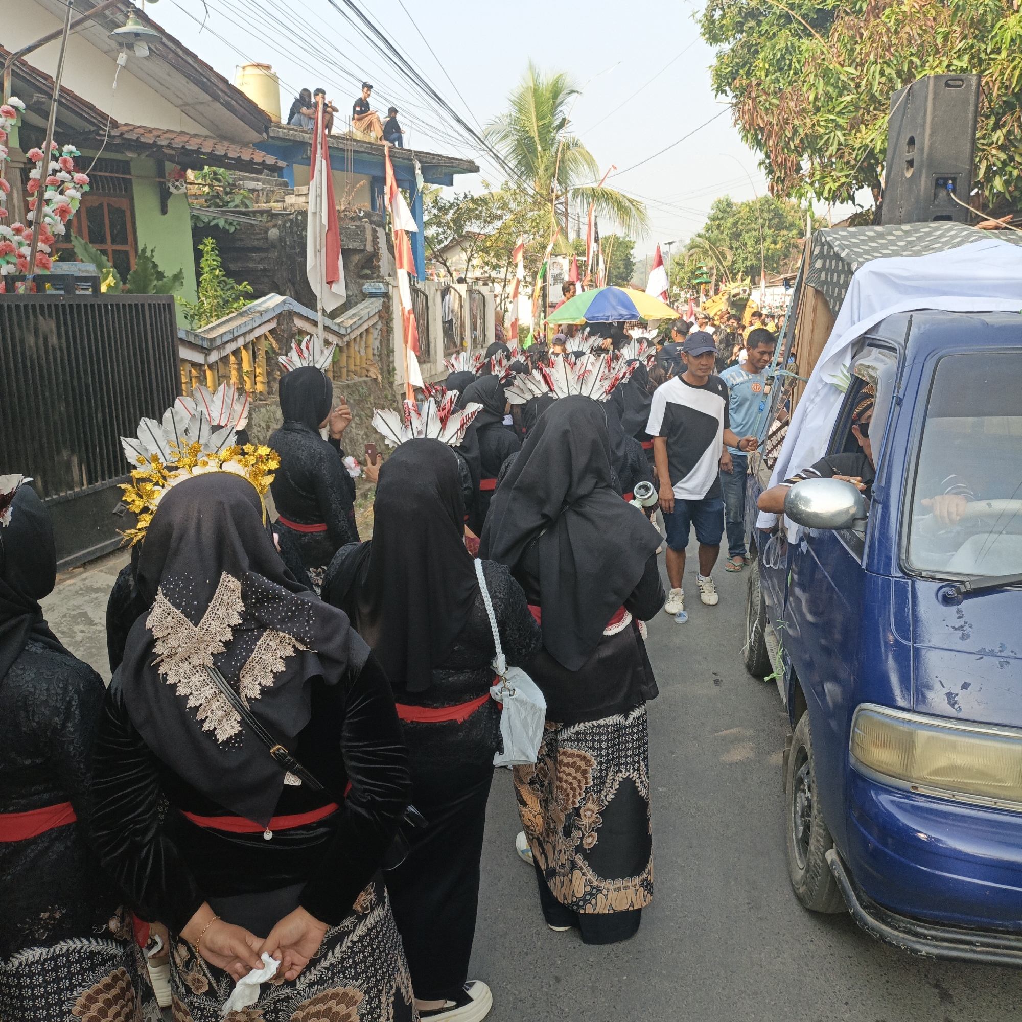 This is a medium shot of a parade or procession on a street in Tanahbaya, Indonesia. In the foreground, several individuals are dressed in traditional black attire with ornate gold and white headpieces featuring feathers. They are walking along the street. To the right, a blue truck is visible, partially covered with a white tarp, with a person in the driver's seat. Other individuals, some in casual clothing and others also in procession attire, are seen further down the street. Indonesian flags are prominently displayed on poles and attached to buildings. The environment appears to be a residential street with houses lining the sides, and lush green trees in the background. The sky is bright and clear, suggesting it is daytime.