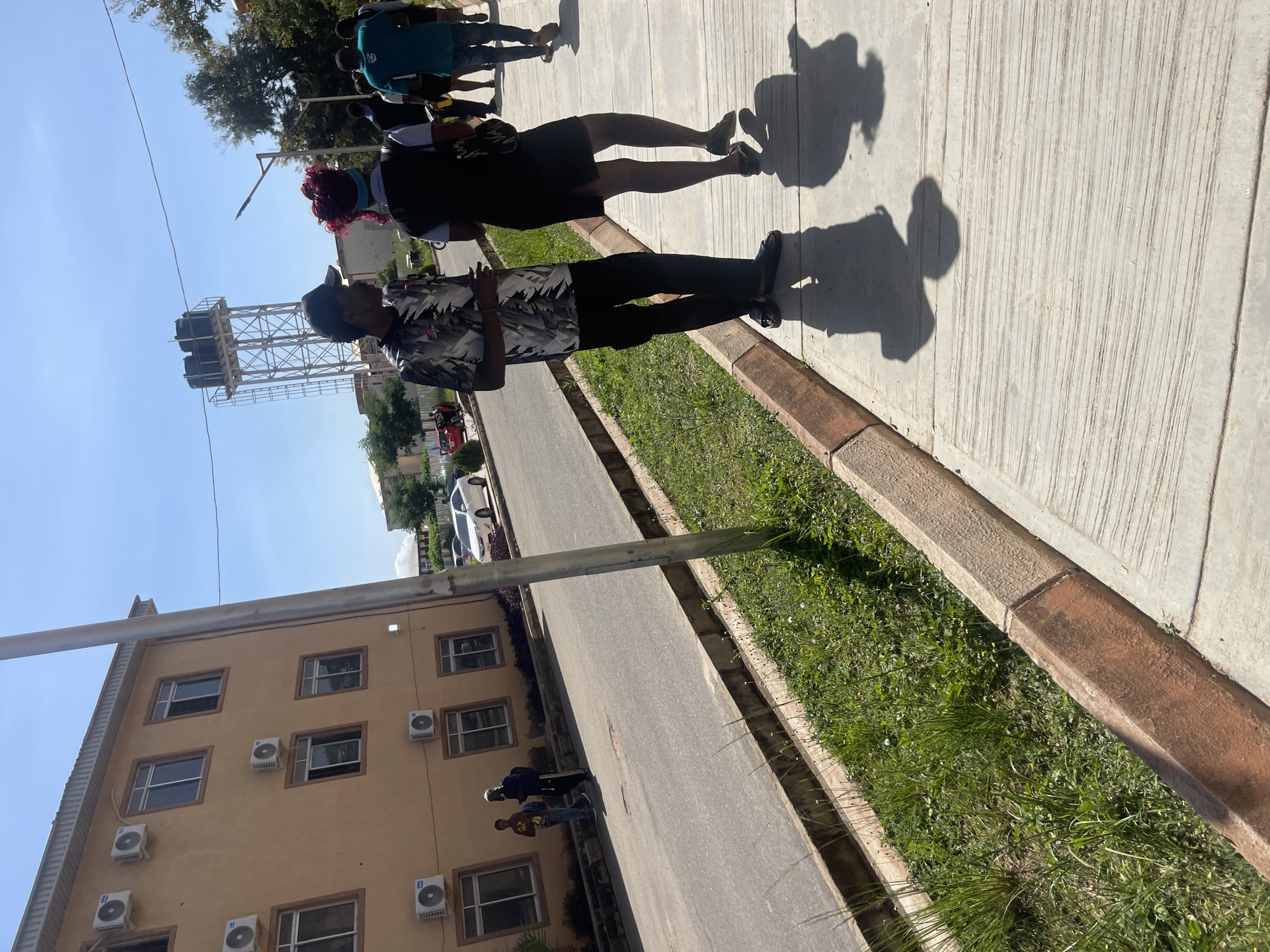 The image depicts a sunny outdoor scene on a paved walkway adjacent to a road, with a building on the left and trees and a water tower in the background. The location is identified as Rafin Par, Nigeria.
In the foreground, two individuals are walking away from the camera on the right side of the walkway. The person in front is wearing a black dress and has bright red hair, with a dark vest over a lighter top. They are carrying a dark bag. The person behind them is a male wearing a patterned short-sleeved shirt, dark pants, and a black baseball cap. They are casting long shadows on the ground, indicating the bright sunlight. Other individuals are visible further down the walkway, walking in the same direction.
The building on the left is a two-story structure with a light orange facade. It has several windows with dark frames and multiple air conditioning units mounted on its exterior walls. A grey pole, possibly a lamppost, stands on the walkway between the building and the road.
The road is asphalt and appears to be a street or access road. There are parked cars visible further down the road, including a white sedan. On the left side of the road, there is a strip of green grass and vegetation, bordered by a concrete curb. In the background, beyond the road, a tall metal structure with two water tanks on top stands against a clear blue sky. Trees and other buildings are also visible in the distance. The overall impression is a bright, daytime scene in a developed area.