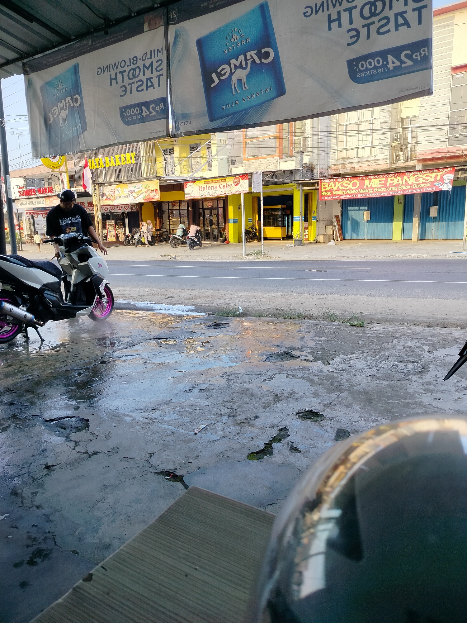 The image depicts a street scene in front of several businesses in Braja Asri, Indonesia. In the foreground, a person is washing a white and black motorcycle, which has pink accents on its wheels. Water and soap suds are visible on the wet, cracked concrete surface. A motorcycle helmet is partially visible in the bottom right corner, reflecting the environment. Behind the motorcycle, several shops are visible, including one with a yellow facade and signage for "JAYA BAKERY." Another banner above the shops advertises "Camel" cigarettes. Further down the street, a red banner announces "MEAT BALL BAKSO MIE PANGS" with an address in "BRAJA SAKTI WAY JEPARA." The street is paved, with parked motorcycles and people in front of the businesses. The overall impression is of a typical commercial street on a bright day, with a focus on daily activities like vehicle maintenance and local commerce.