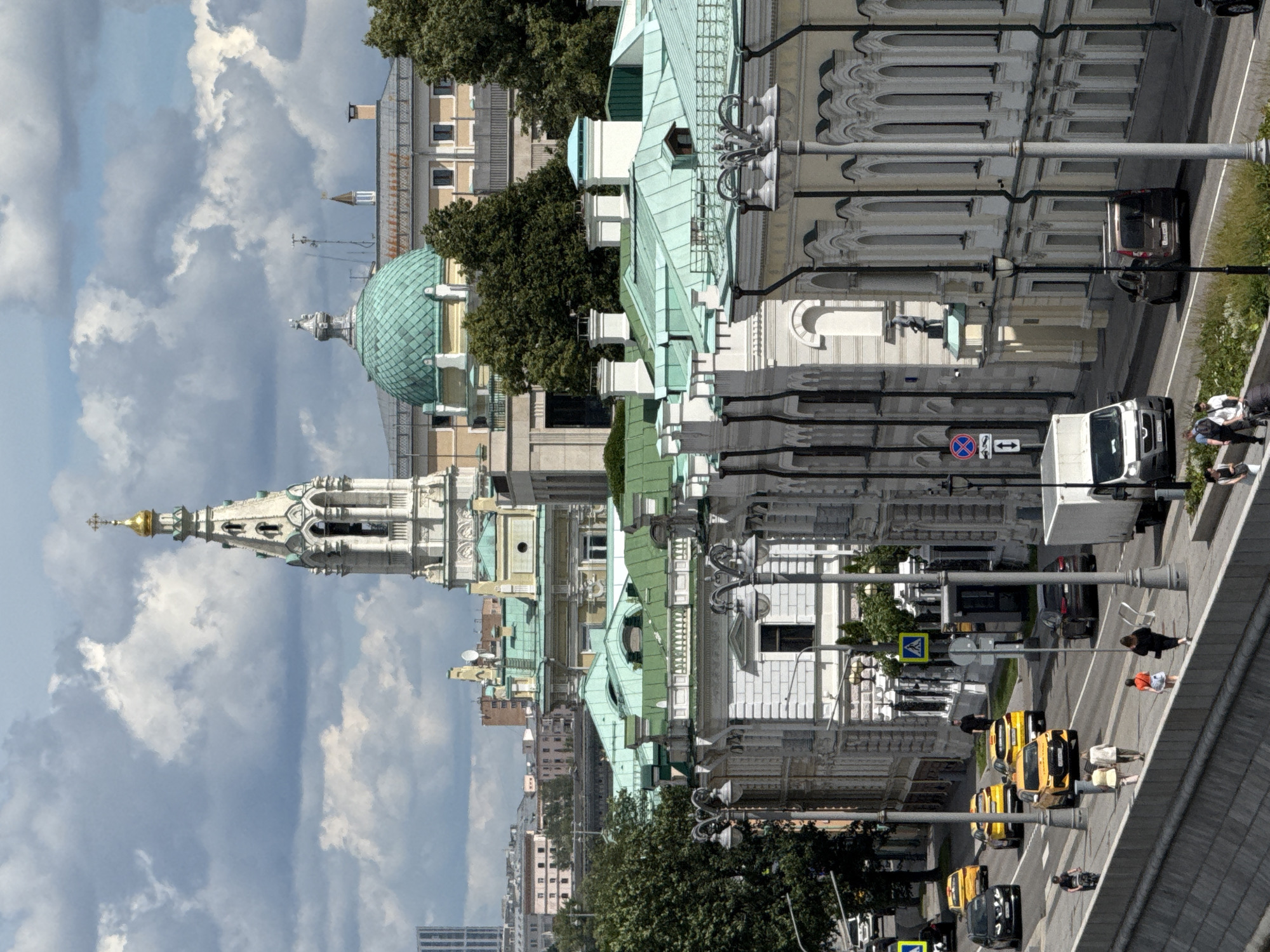 This image depicts a street scene in Moscow, Russia, with a mix of historical and modern architecture. The perspective is from an elevated viewpoint, looking down at the street and buildings. The sky is filled with dramatic, heavy clouds, suggesting potentially overcast or stormy weather.
Prominently featured are several ornate buildings with green roofs and classical architectural elements, including domes and detailed facades. One building has a pointed spire topped with a golden cross. In the foreground, a street runs alongside these buildings, with several yellow taxis and a white truck visible. Pedestrians are seen on the sidewalk. The scene appears to be during daylight hours, though the cloudy sky may be obscuring direct sunlight.
Traffic is moving along the street, with the taxis appearing to be waiting or driving. There are traffic signs visible, including a blue circular sign with a red border and a diagonal red line, possibly indicating a "no entry" or "no stopping" restriction, and a yellow diamond-shaped sign with a pedestrian crossing symbol. The overall impression is a busy urban environment. The people in the image are too small to discern specific emotions or activities beyond walking or standing. There is no discernible text in the image, except for what might be on license plates of the vehicles.
