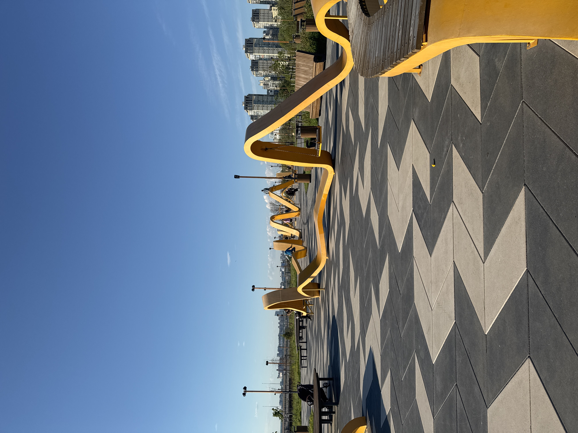 The image depicts a public space in Saint Petersburg, Russia, characterized by modern urban design. A prominent yellow, ribbon-like structure serves as seating, winding through the paved area. The pavement is composed of dark gray and light gray chevron-patterned tiles. In the background, multi-story buildings are visible, suggesting a residential or commercial district. Several people are seated on the yellow structure. Streetlights are spaced along the walkway. The sky is clear blue. Benches are also visible along the walkway.