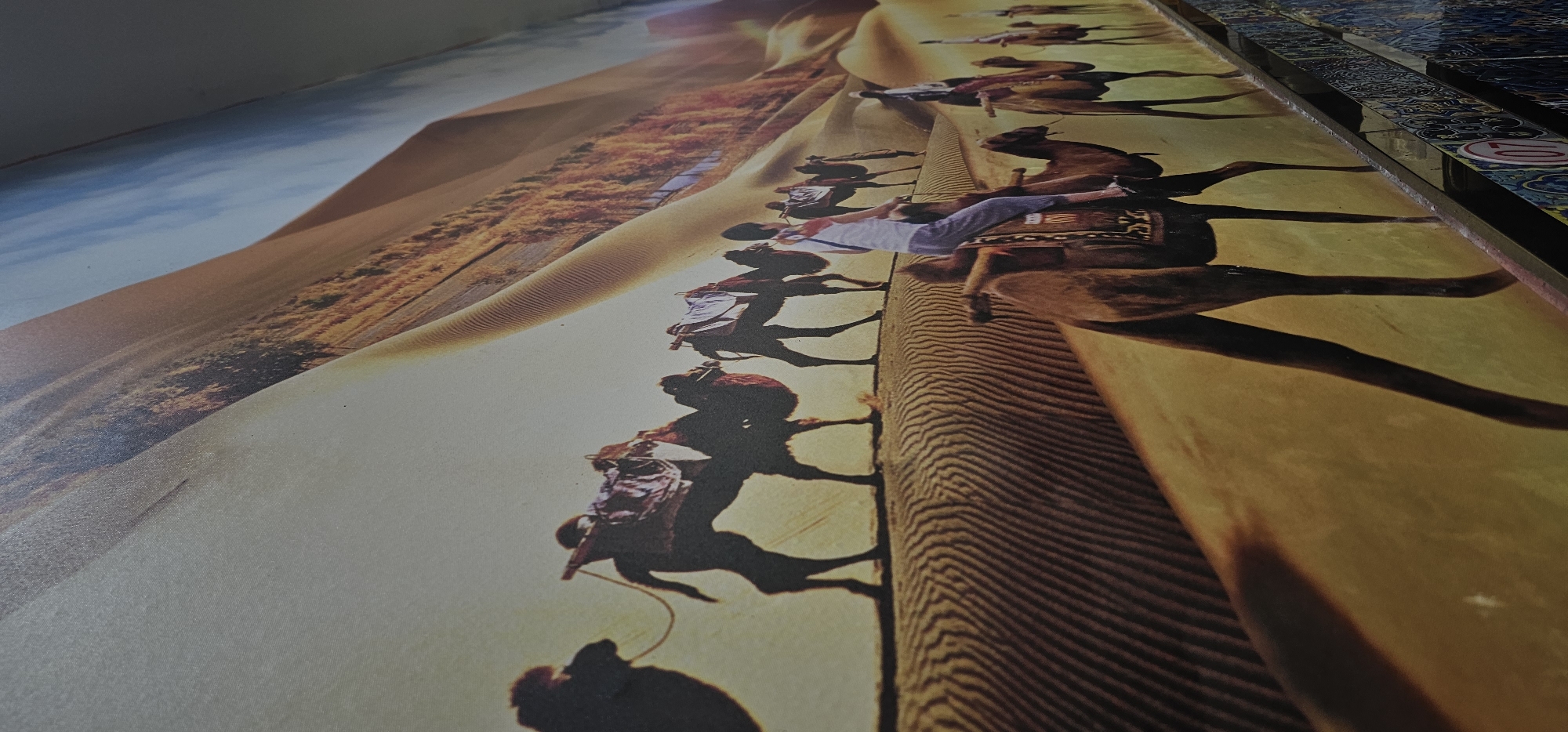 A slightly angled, close-up shot shows a print depicting a camel caravan traversing a desert landscape, likely set within Yuzhong District, China. The print, mounted on a surface adjacent to decorative tiles, captures a line of camels with riders against a backdrop of sand dunes and a blue sky. The lighting is uneven, casting shadows and highlighting textures within the print. The focus is sharpest on the central portion of the caravan.
