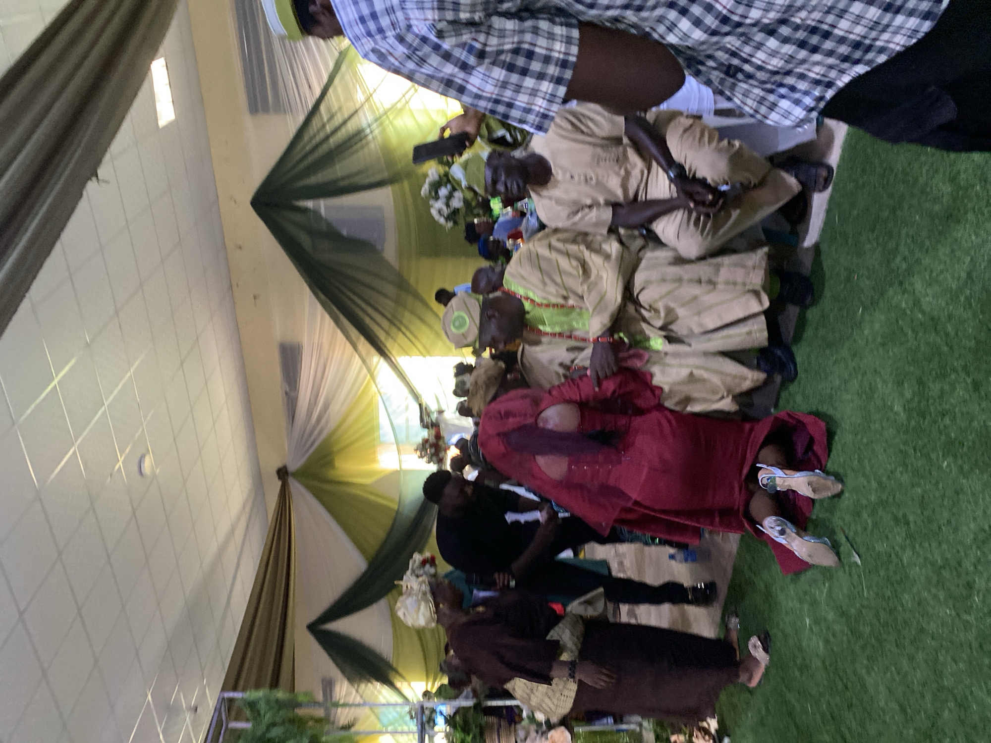 The image depicts a group of people, likely attending an event, possibly a wedding or celebration, based on their attire and the decorations. The location appears to be indoors, with decorative fabric draping from the ceiling, featuring colors such as green, yellow, and white.
Several individuals are seated, wearing traditional clothing, including tan-colored garments. Other individuals are standing, dressed in darker attire. A woman wears a red outfit.
There is artificial green turf on the floor. The location is stated to be in Kuru Karama, Nigeria.
