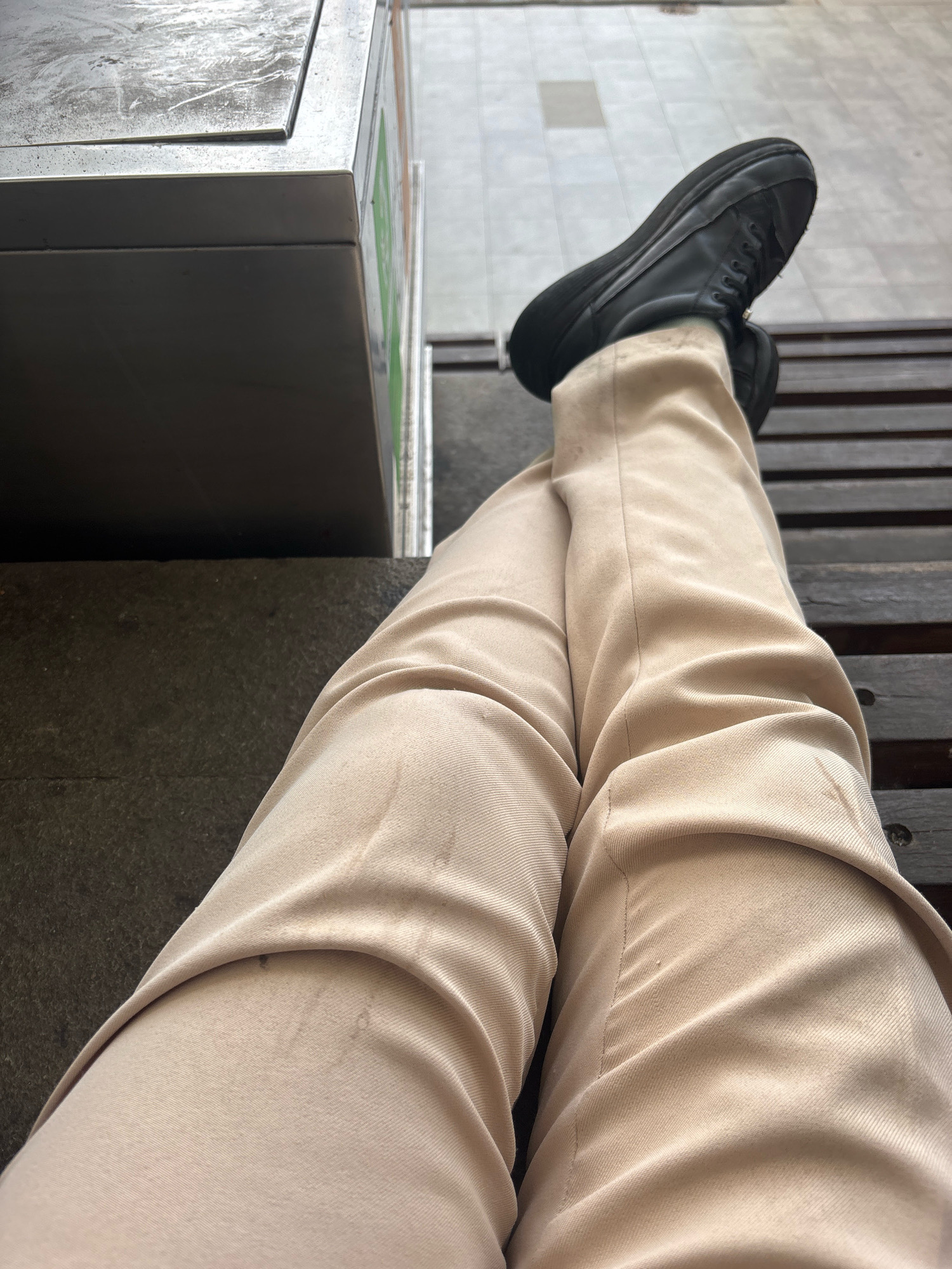 A person is sitting on a wooden bench with their legs crossed. They are wearing light-colored pants and black sneakers. To the left of the person is a metal structure with a textured surface. The environment appears to be outdoors, possibly in a public area with a tiled floor in the background. The scene is captured from a high-angle perspective, focusing on the person's legs and the immediate surroundings. The location is specified as North Jakarta, Indonesia.