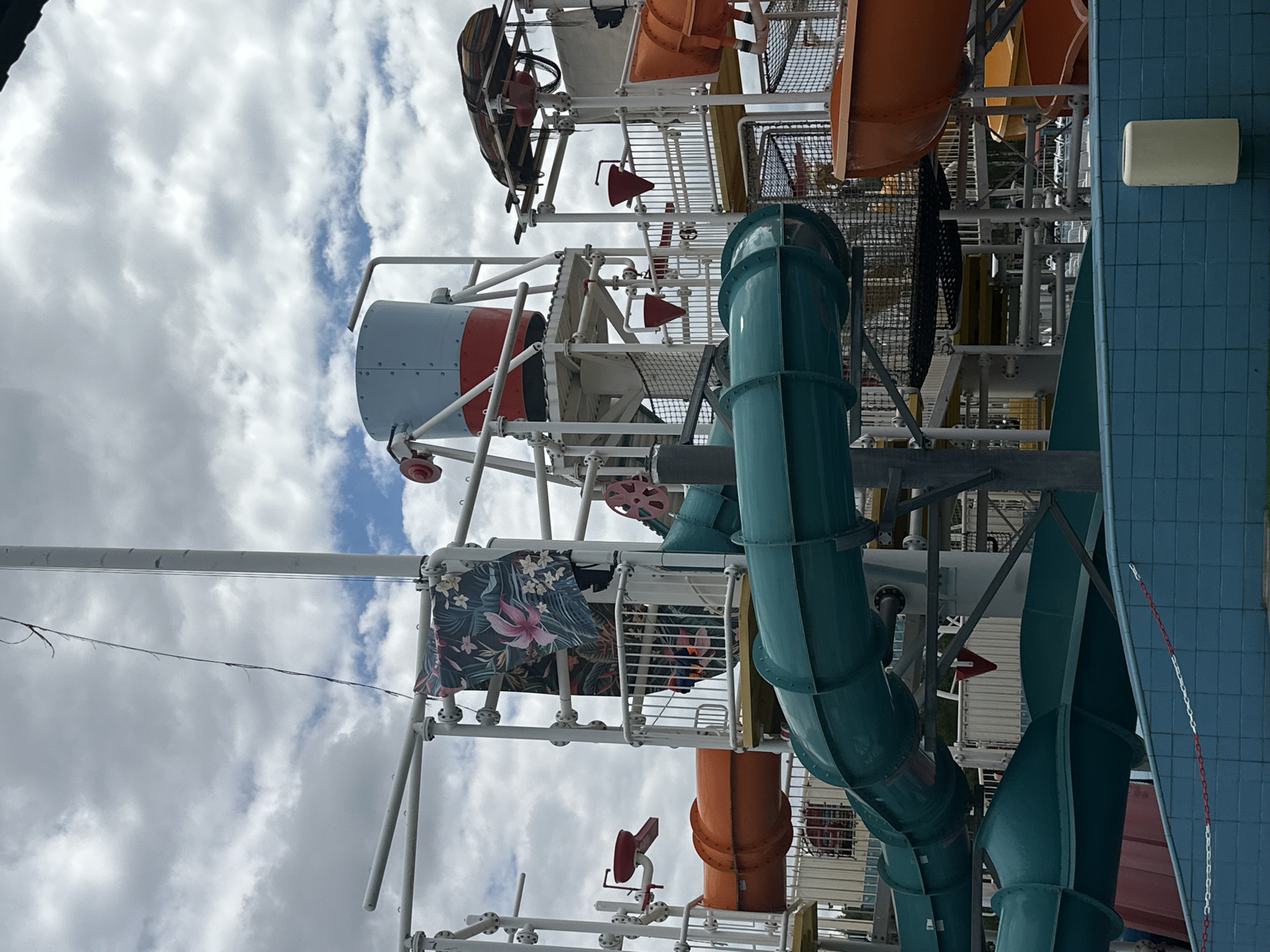 The image depicts a section of a water park's water slides, set against a cloudy sky. The structure is made of white metal supports with various colored slides, predominantly teal and orange. There is a large, light blue and red bucket-like structure at the top of one of the platforms, suggesting a tipping bucket water feature. A floral-patterned fabric hangs from a railing. In the foreground, a blue tiled pool edge is visible, with a red and white chain barrier. The overall scene suggests an outdoor recreational facility. There are no people visible in the image. The location is not identifiable as Minsk, Belarus.