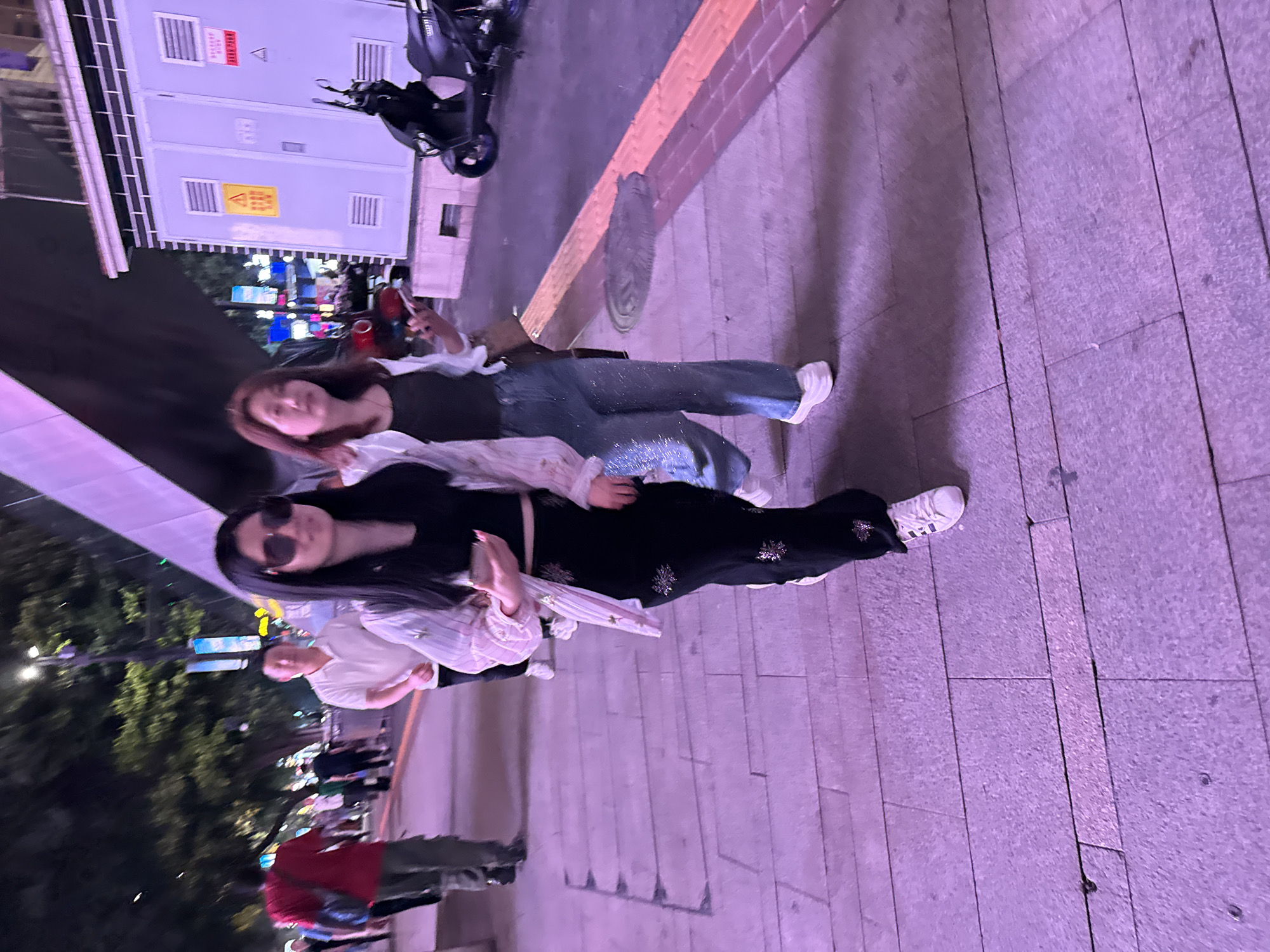 The image, rotated counter-clockwise, captures a street scene in what appears to be an urban setting, likely in Yuzhong District, China. Two young women are standing on a sidewalk paved with rectangular tiles. The woman in the foreground has long, dark hair and is wearing sunglasses, a black crop top, black pants with floral embellishments, a light-colored striped jacket, and white sneakers. She is holding what appears to be a plastic bag or container. The woman behind her has shoulder-length brown hair and is wearing a dark top, blue jeans with embellishments, a light-colored jacket, and white shoes. In the background, there is a building with signs in Chinese and English, a parked scooter, trees, and indistinct figures of people walking. The light suggests it is nighttime.