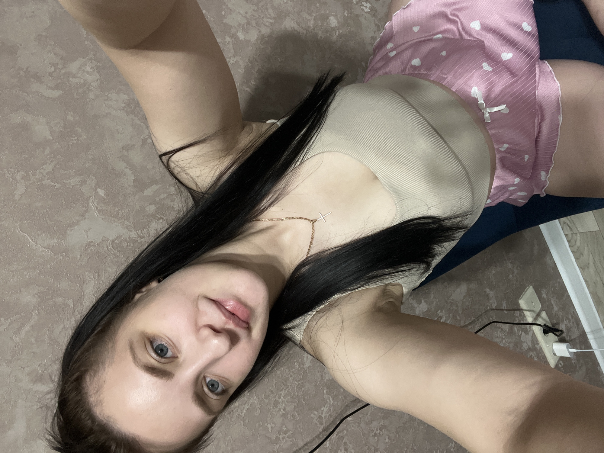 A selfie of a woman in Марковский, Russia, wearing a beige ribbed tank top, pink and white heart-printed shorts, and a gold necklace with a cross pendant, lying on a patterned surface. Her dark hair is long and partially covers her chest. A blue surface is visible behind her waist. A power outlet with a charger plugged in is on the wall behind her legs.