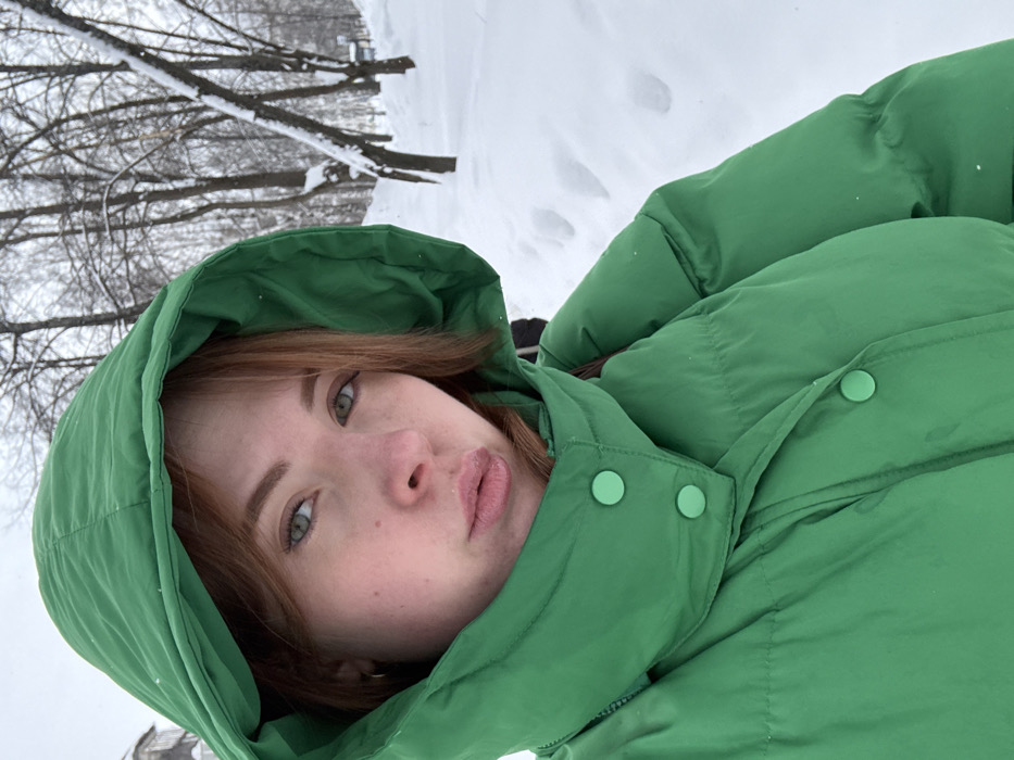The image depicts a fair-skinned female individual in her 20s or early 30s taking a selfie in a snowy outdoor environment within the city of Chaikovsky, Russia. She is centrally positioned, looking directly at the camera with light-colored eyes and reddish-brown hair partially visible beneath a bright green hooded puffer jacket. The jacket features two prominent light green snap buttons visible on the high collar. Her lips are slightly pursed, and her face shows no discernible injuries or anomalies. The background consists of a snow-covered landscape with several bare deciduous trees dusted with snow. Footprints are visible in the deep snow on the ground. A light-colored building or structure is partially visible in the upper left background. The scene suggests an overcast winter day.