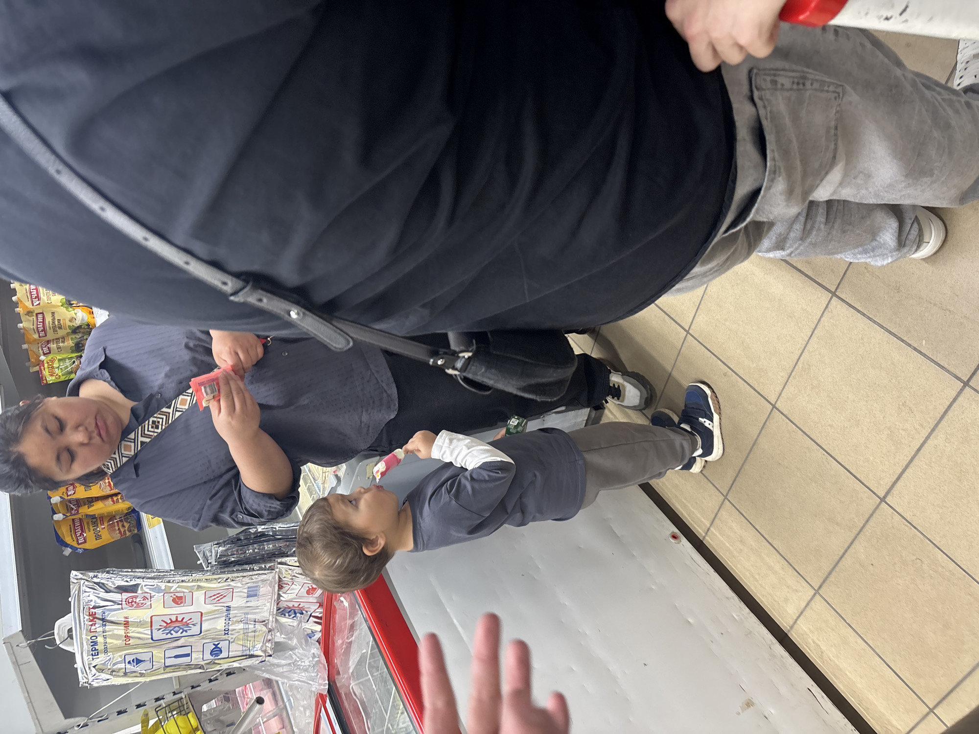 In a retail environment, a young boy with brown hair stands to the left, eating a pink popsicle. He is wearing a dark grey t-shirt over a white long-sleeved shirt and grey sweatpants, with blue and white sneakers. A woman, likely his mother, stands behind and to his right, looking down at something in her hands. She has dark hair pulled back and is wearing a dark blue collared shirt with a patterned strap across her chest. Partially visible in the foreground is the back of another person wearing a black t-shirt and light grey sweatpants. The background shows shelves stocked with various products, including packaging for thermal bags and food items. The floor is tiled in a beige, grid pattern, and a refrigerated display case is visible to the left of the boy. The scene appears to be a grocery store or supermarket in an unknown city in Russia.