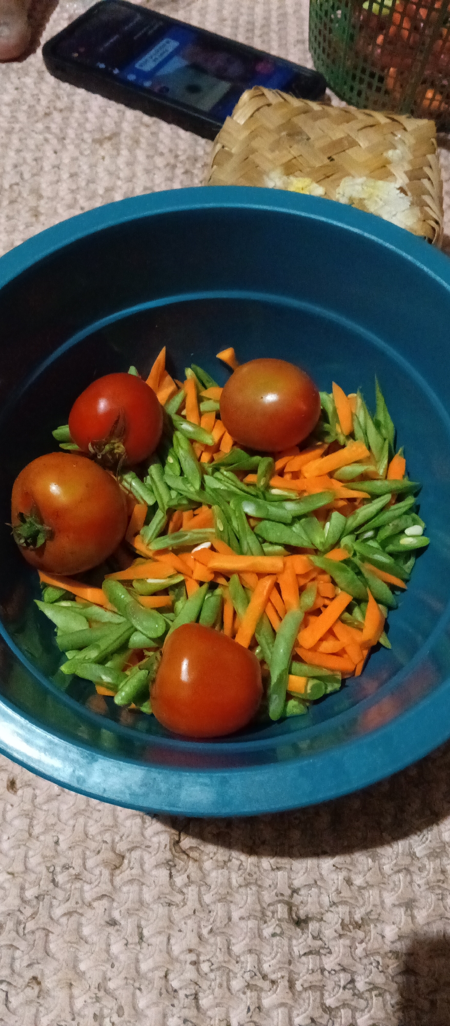 In Sukahurip, Indonesia, a close-up shot captures a blue bowl filled with chopped vegetables and whole tomatoes. The bowl sits on a textured beige surface. Inside the bowl, a colorful mix of vibrant orange carrot pieces and slender green beans is visible. Four ripe red tomatoes are interspersed with the vegetables. In the blurred background, a smartphone displays a screen with text, and a woven bamboo container with a light-colored filling is also present. The lighting suggests an indoor setting, possibly a kitchen or dining area.