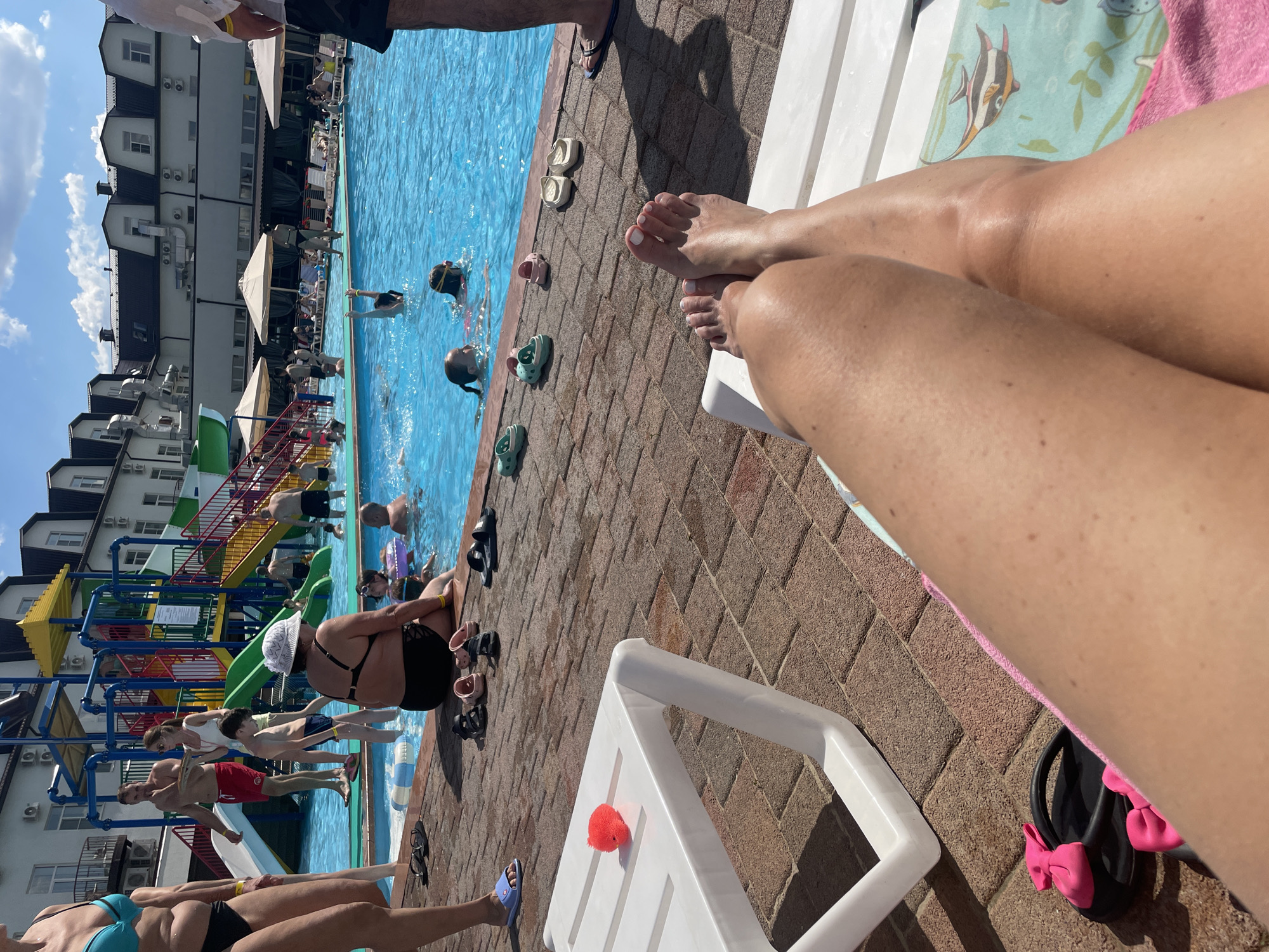 In Пристанное, Russia, a sunny day at a water park features a blue swimming pool, a colorful water slide structure, and a white hotel building. Several people are visible, including adults and children, some swimming or interacting in the pool, while others relax on lounge chairs or walk around the poolside. On the brick-paved area, various footwear like sandals, flip-flops, and Crocs are scattered. A white plastic sun lounger in the foreground displays a pink fuzzy object and a towel with a fish pattern.