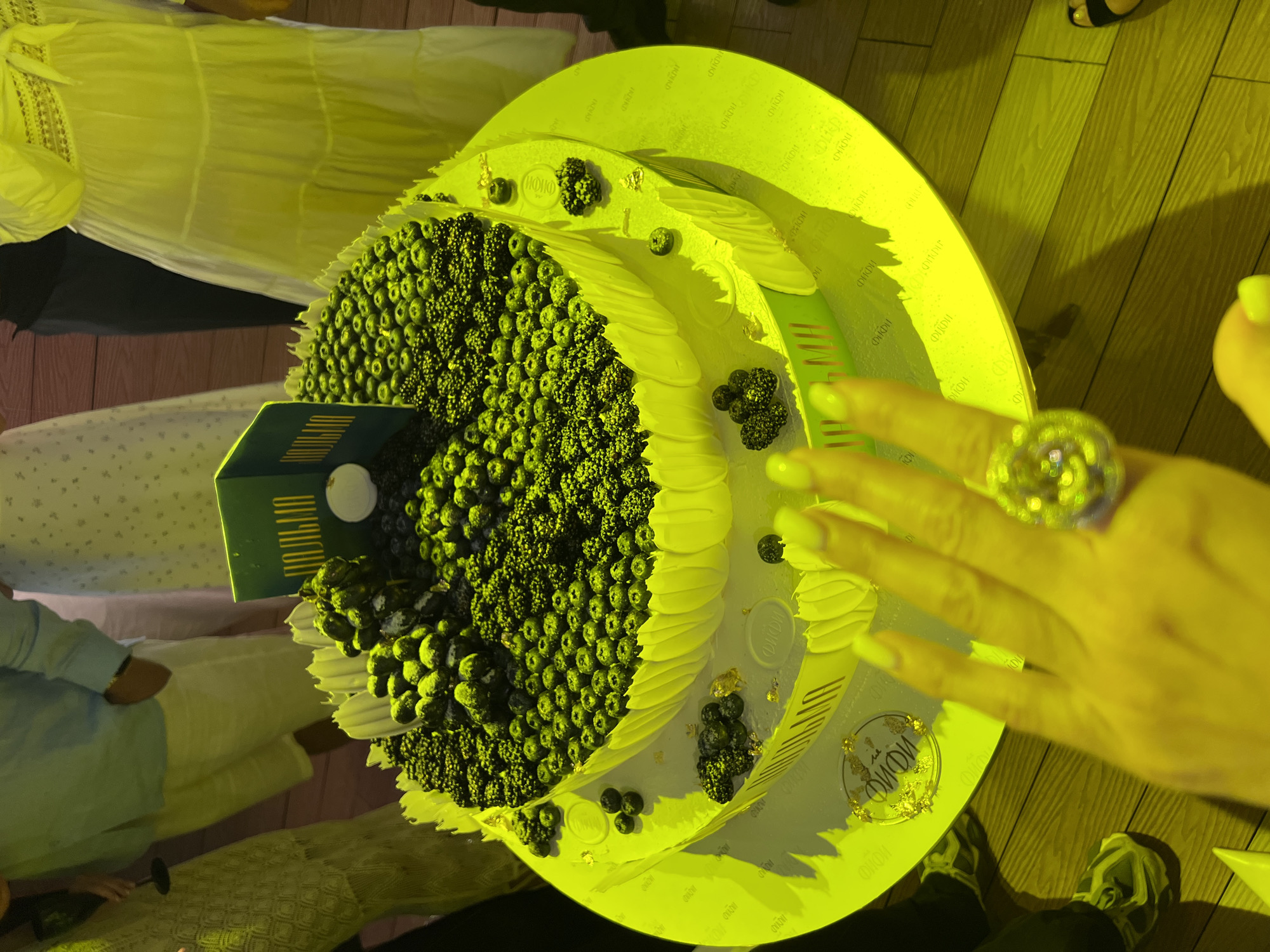 The image shows a two-tiered cake covered in blueberries and blackberries. A green sign with Cyrillic writing is placed on top of the cake. The cake is on a round platter, and the platter is on a wooden table. A woman's hand with a large ring is in the foreground, reaching towards the cake. Other people are visible in the background, out of focus, wearing light-colored clothing. The overall lighting is a strong yellow cast, possibly from artificial lights or a filter. The context suggests a celebration or event. It is impossible to determine the specific location in Sochi, Russia, from the provided image.